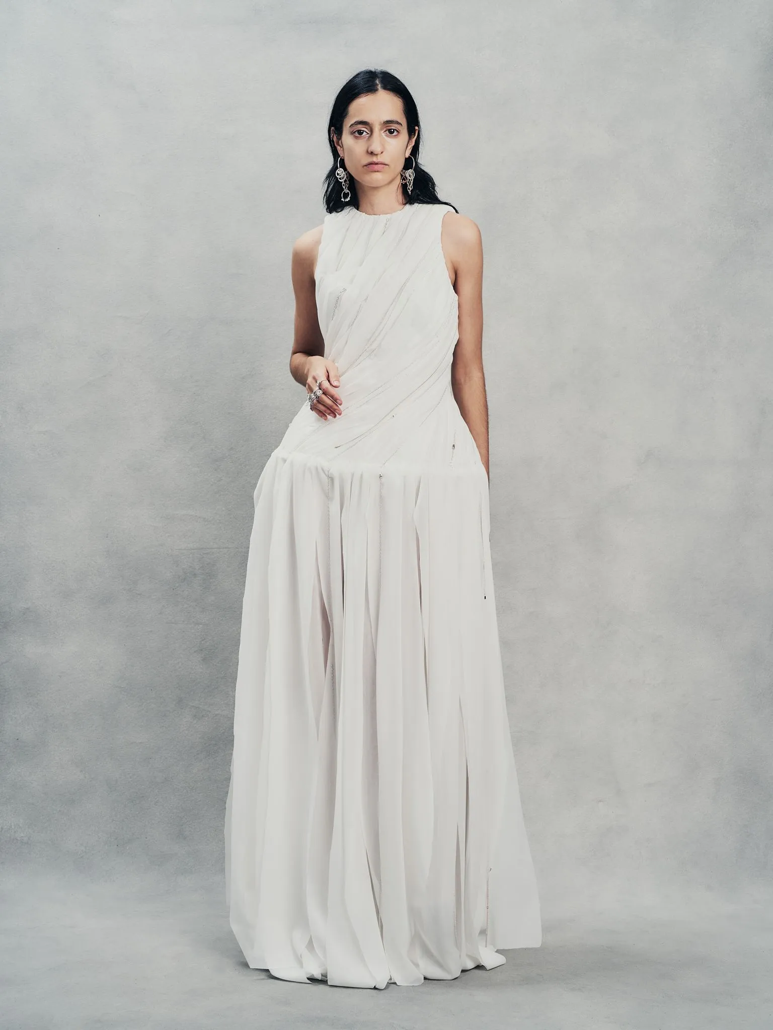 Woman in a white flowing gown standing against a gray backdrop, facing the camera with a neutral expression, wearing large earrings