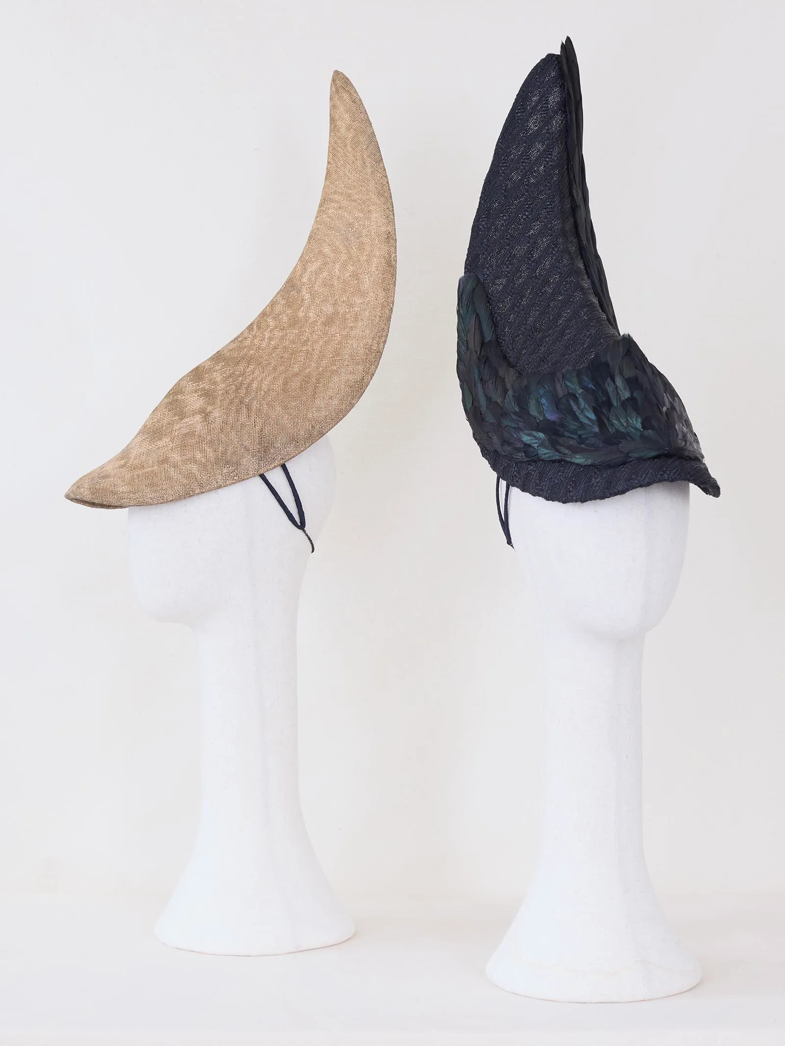 Two horn-shaped hats on white mannequin heads: beige burlap left and dark navy with feathered trim on the right.