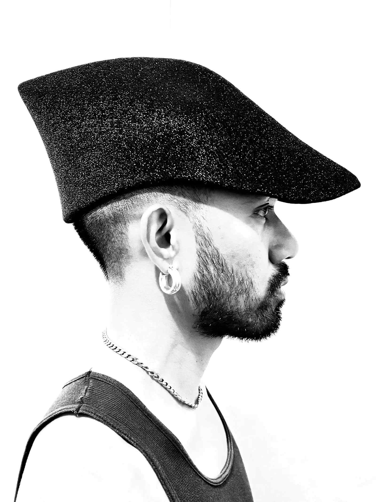 Side profile of a man wearing a glittery black cap, with a short beard, an earring, and a chain necklace visible against a white background.