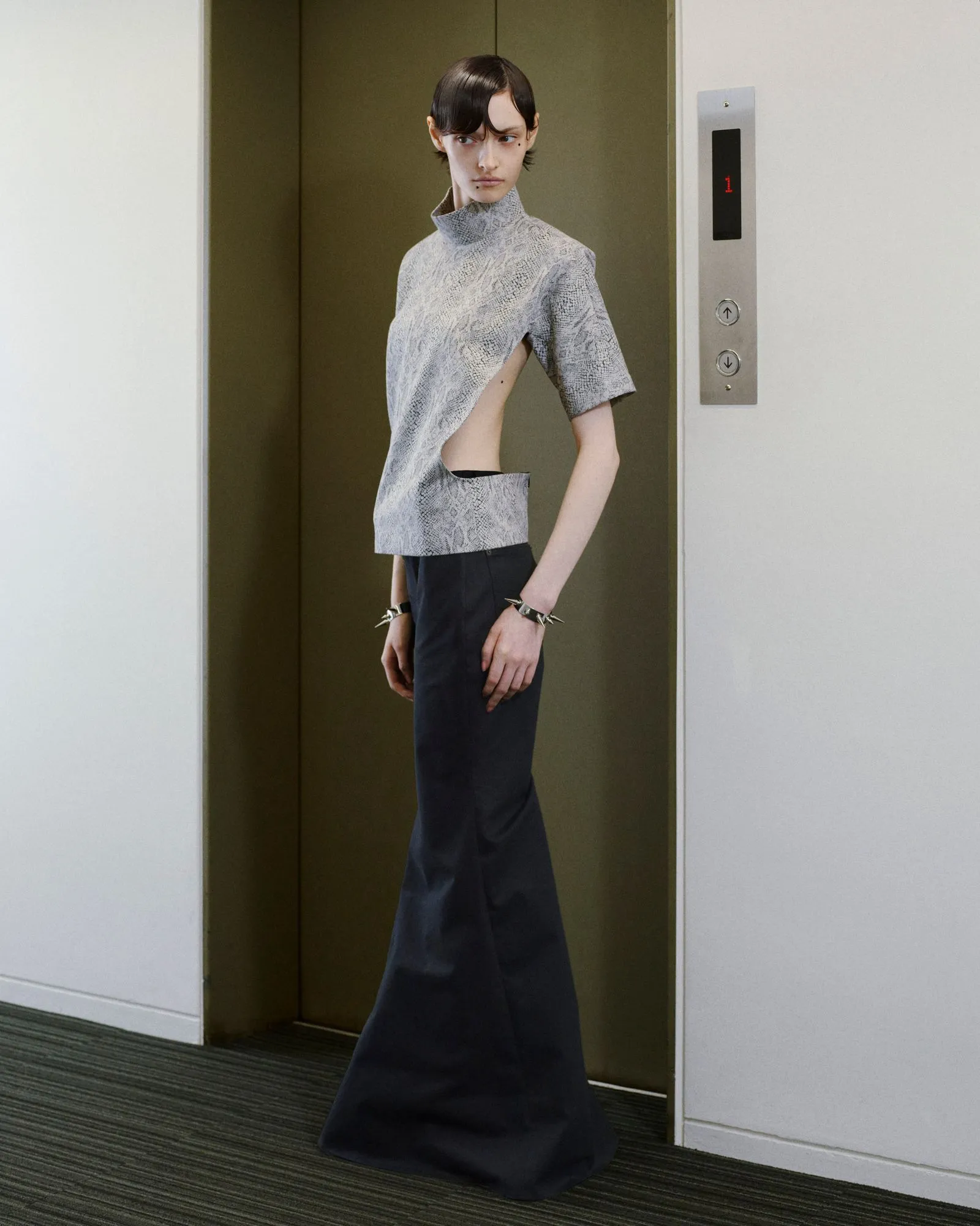 Model standing in an open elevator doorway wearing a gray snakeskin top with side cutouts and dark wide-leg pants, accessorized with spiked bracelets.