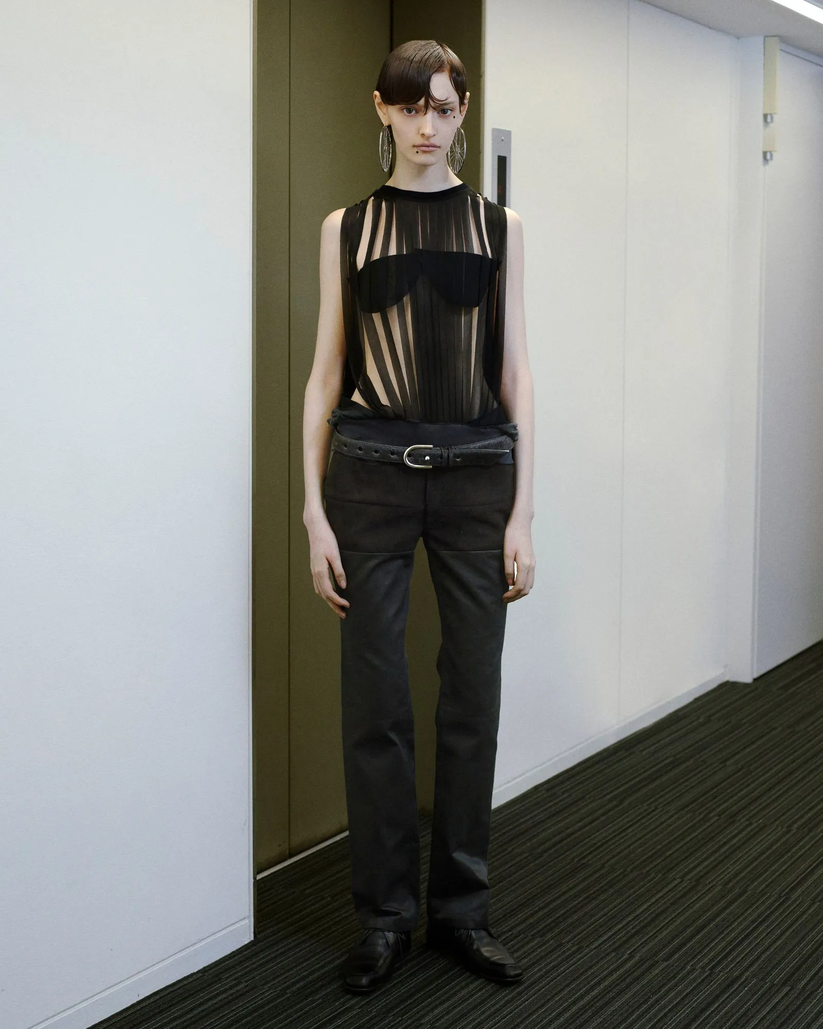 Full-body image of a woman standing in a hallway wearing a sheer black striped top over a black bandeau, black pants with a belt, and large hoop earrings.