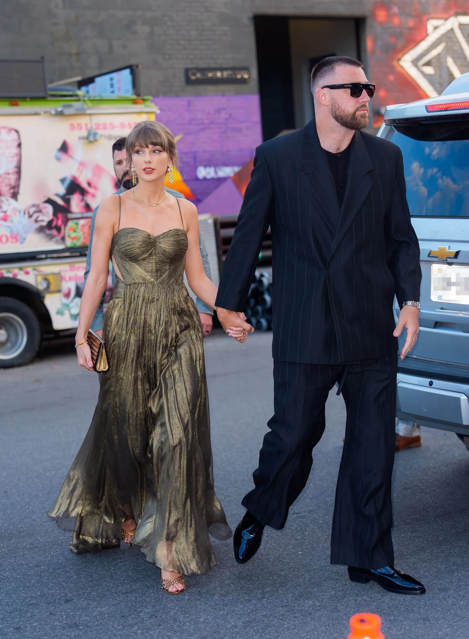 A stylish couple walks hand in hand down a city street; the woman wears a gold metallic gown and heels, the man in a dark pinstripe suit and sunglasses.