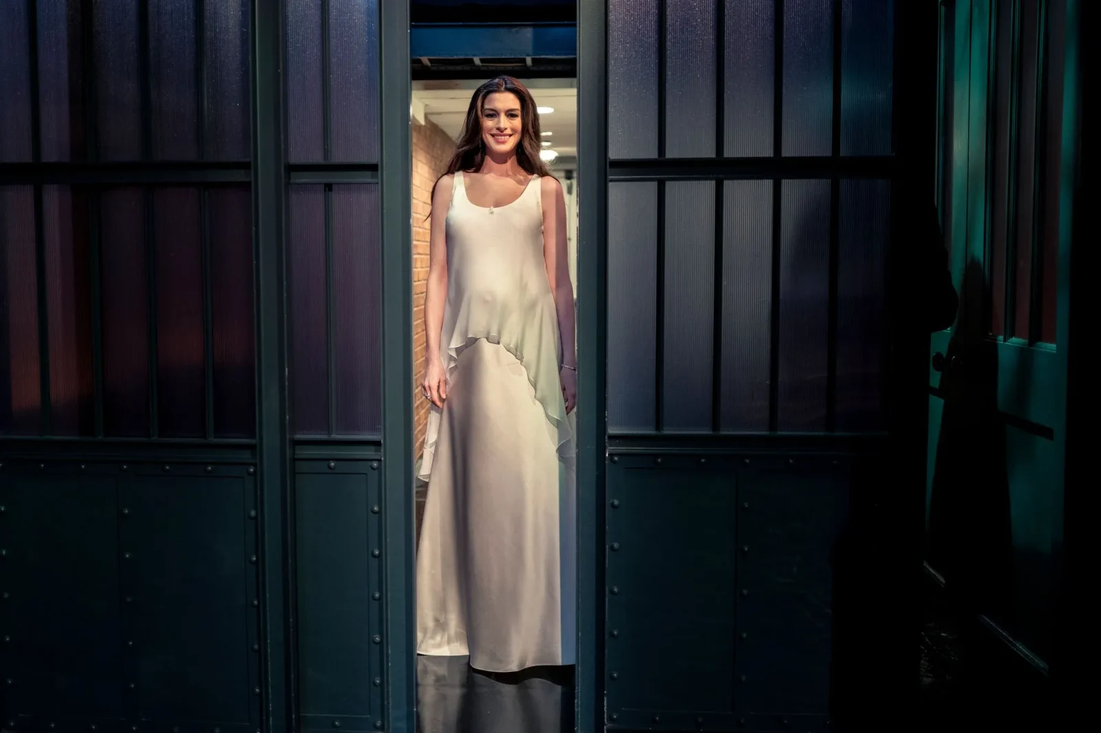 Woman in a white satin evening dress standing in a narrow door frame between dark industrial panels, smiling at the camera.