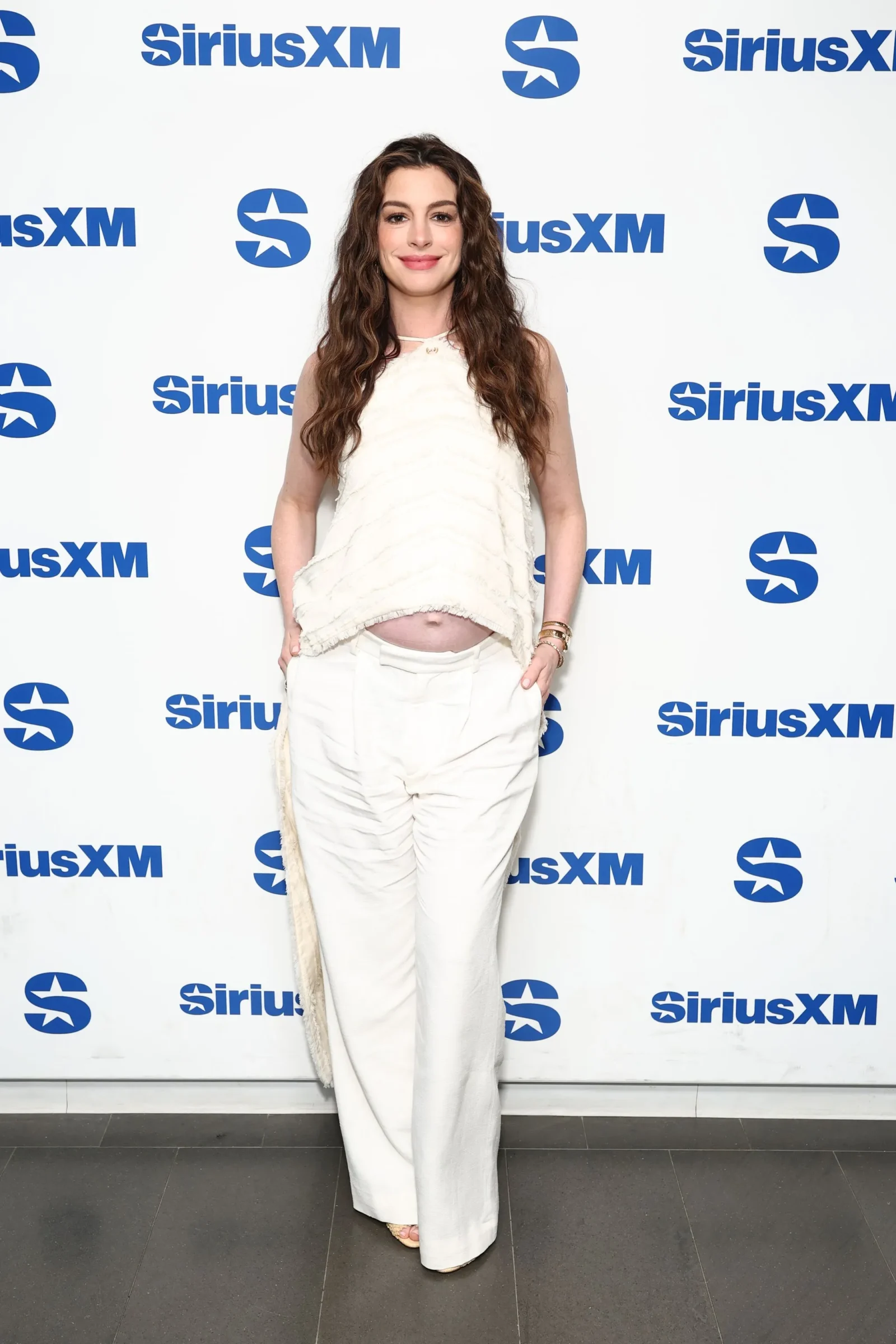 Woman with long wavy brown hair wearing a white sleeveless top and white pants, posing in front of a SiriusXM backdrop with blue logos.