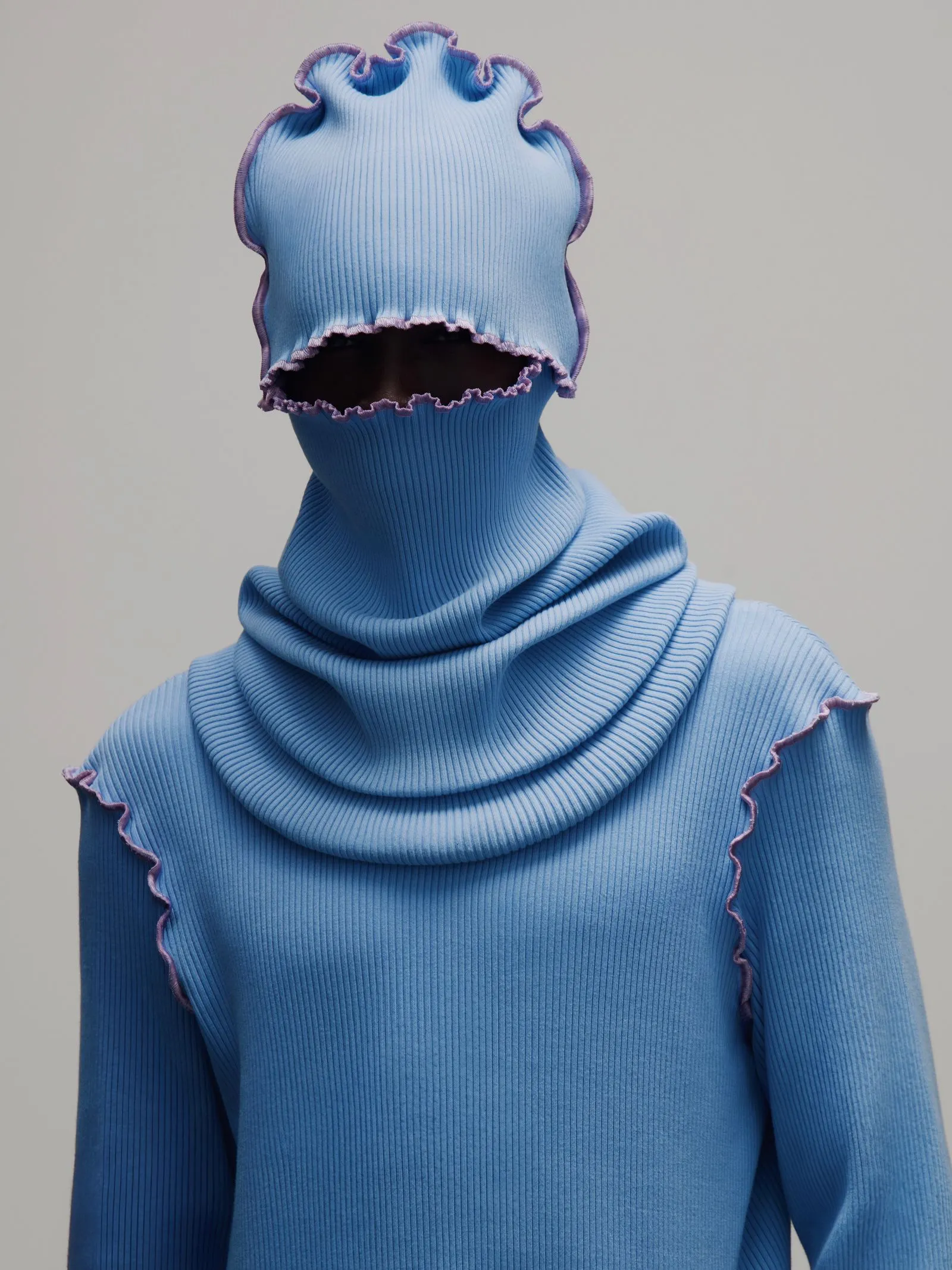 Person in a blue ribbed balaclava with purple ruffled edges covering most of the face, neutral background