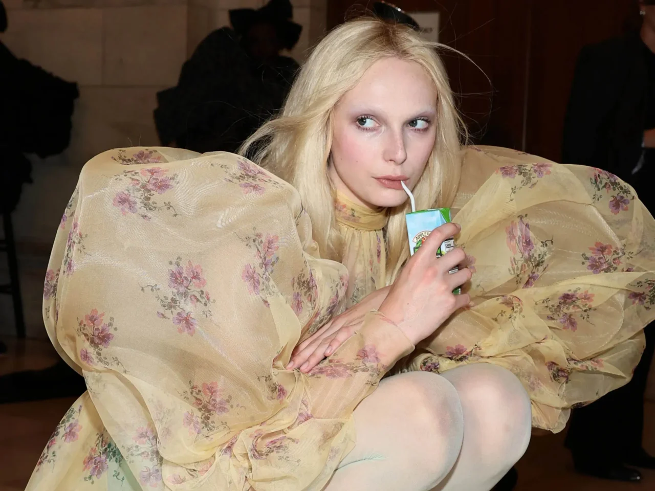 Blonde woman in a sheer floral dress sipping from a small juice box with a straw.
