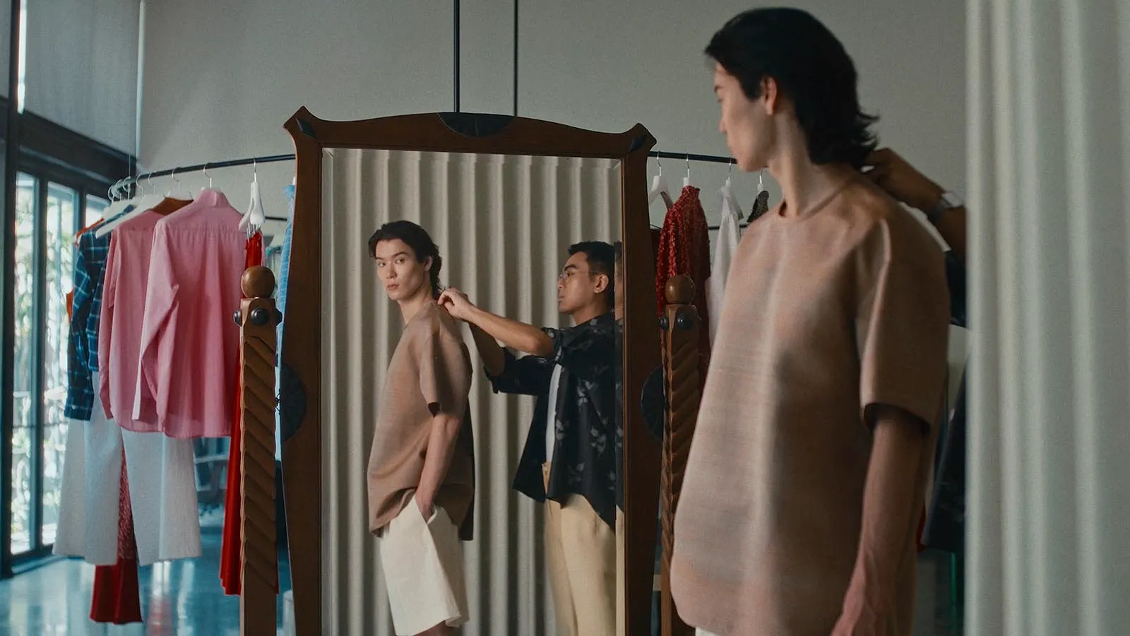 Model wearing beige top standing before a tall mirror while a stylist adjusts the collar in a clothing studio with racks of garments nearby