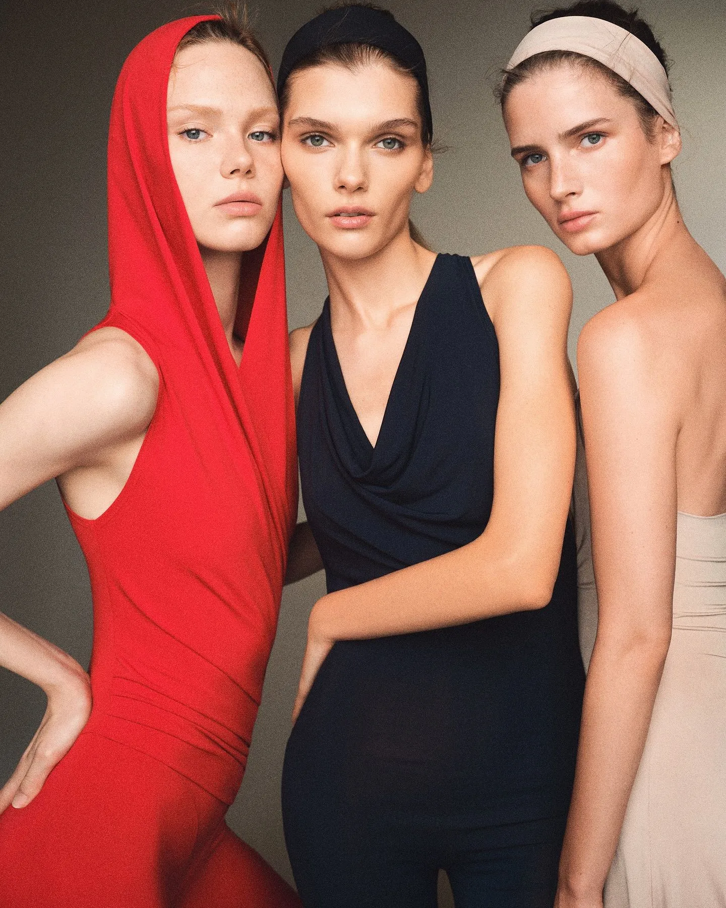 Three fashion models posing closely in red, black, and beige outfits with headbands against a neutral backdrop, showcasing a sleek editorial look.