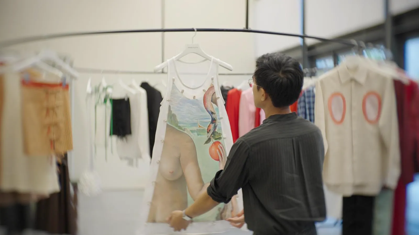 Man browsing a clothing rack in a store, inspecting a white dress with a surreal nude-torso and landscape print on the front.