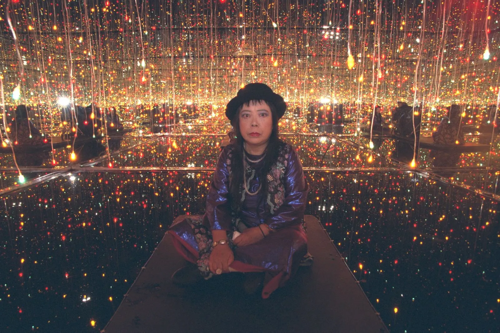 Woman seated on a platform in a mirrored light-filled room with thousands of hanging colorful lights reflection-surrounding her.