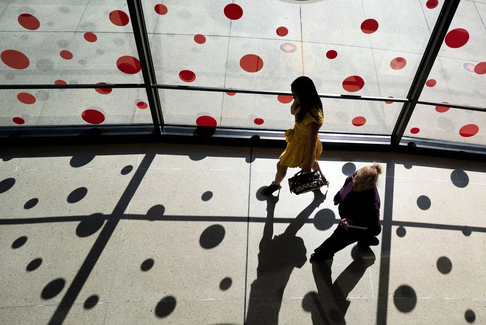 Two people walk side by side in a glass-walled atrium with red polka-dot decals, casting long shadows on the floor.