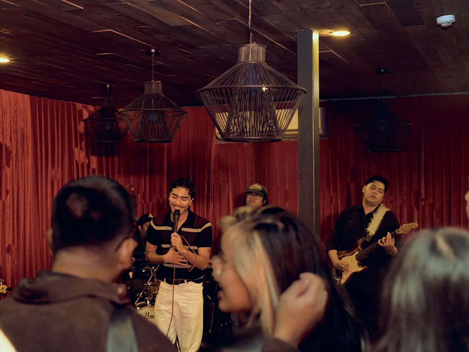 Male vocalist sings into a microphone on a small stage, with a guitarist in the background and red curtains behind them.