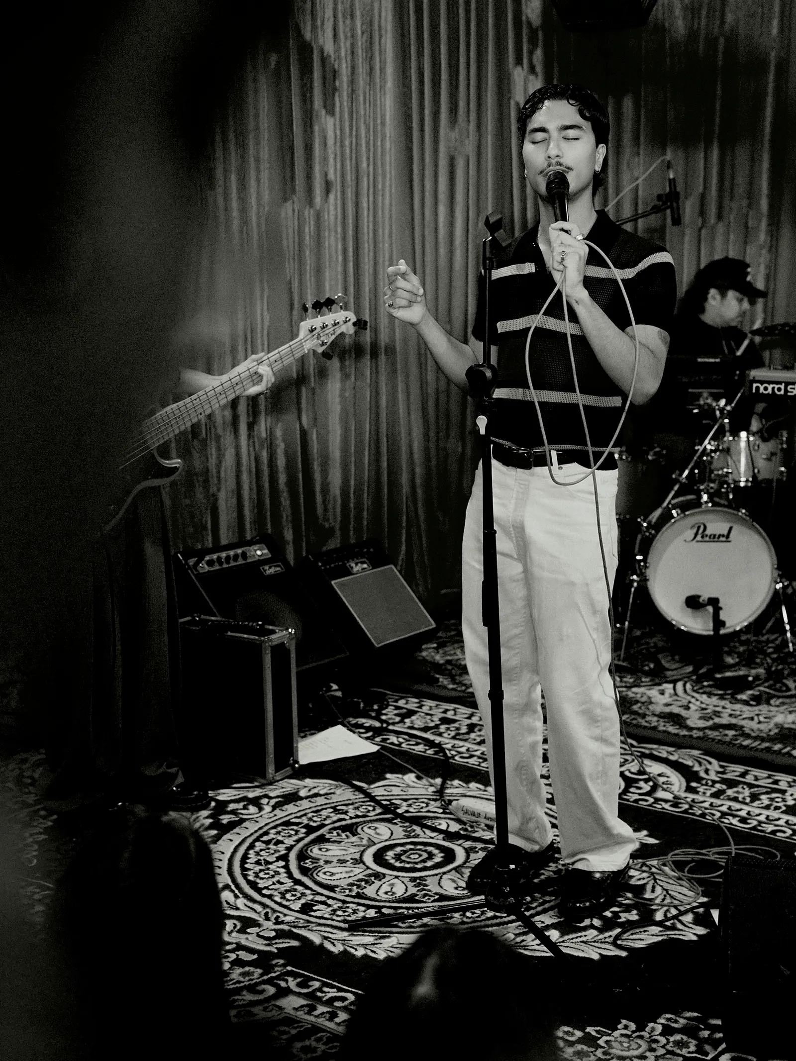 Male singer with eyes closed at a microphone on stage, guitar and drummer visible behind him.