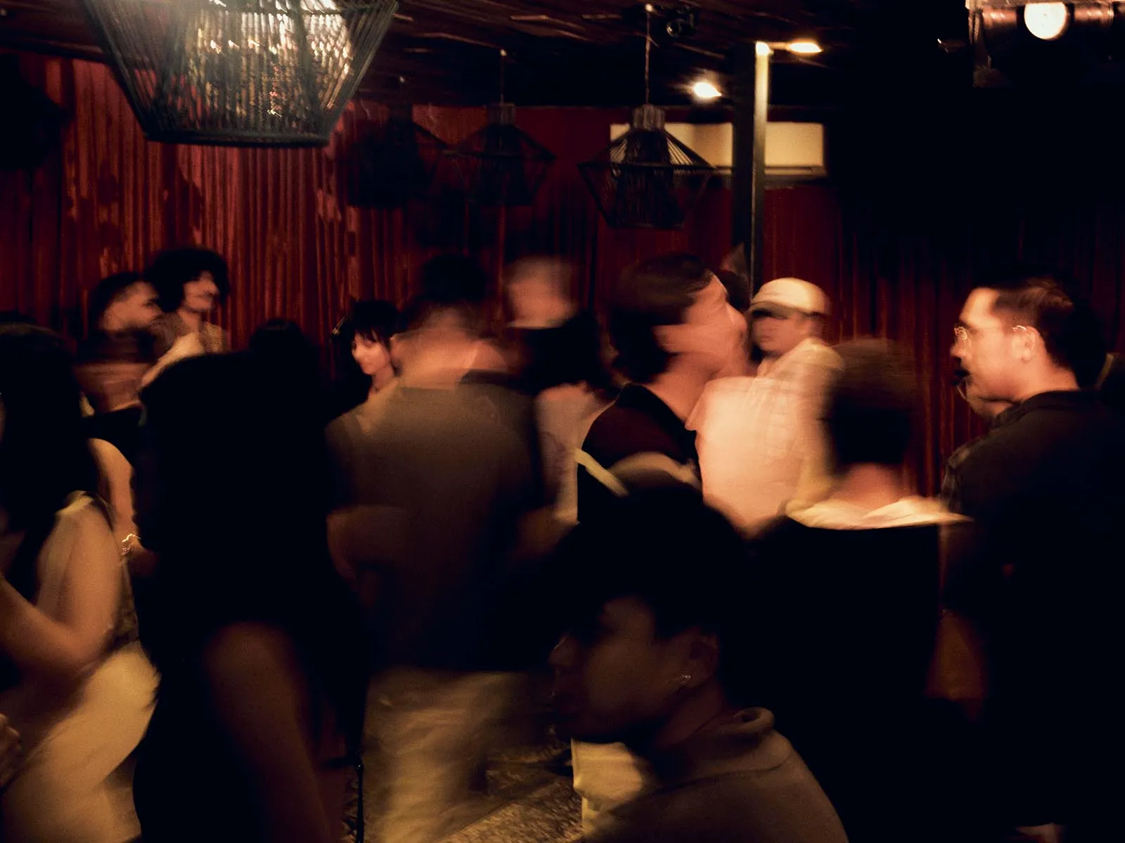 Crowded dance floor in a dimly lit club with red curtains and warm overhead lights, motion-blurred dancers.