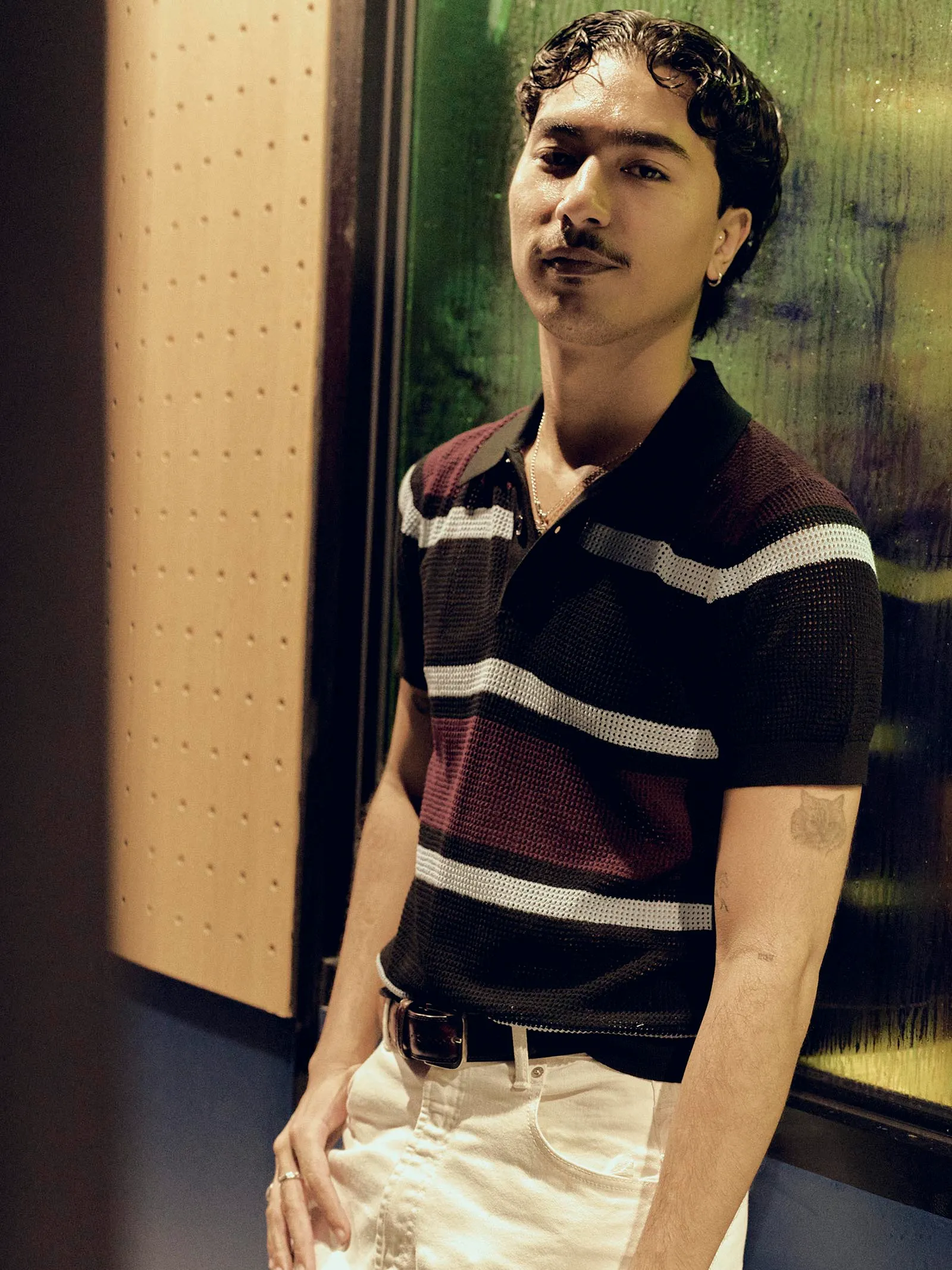 Young man wearing a maroon, white, and black striped polo shirt stands in a dim subway car, leaning against the wall with hands at his side.