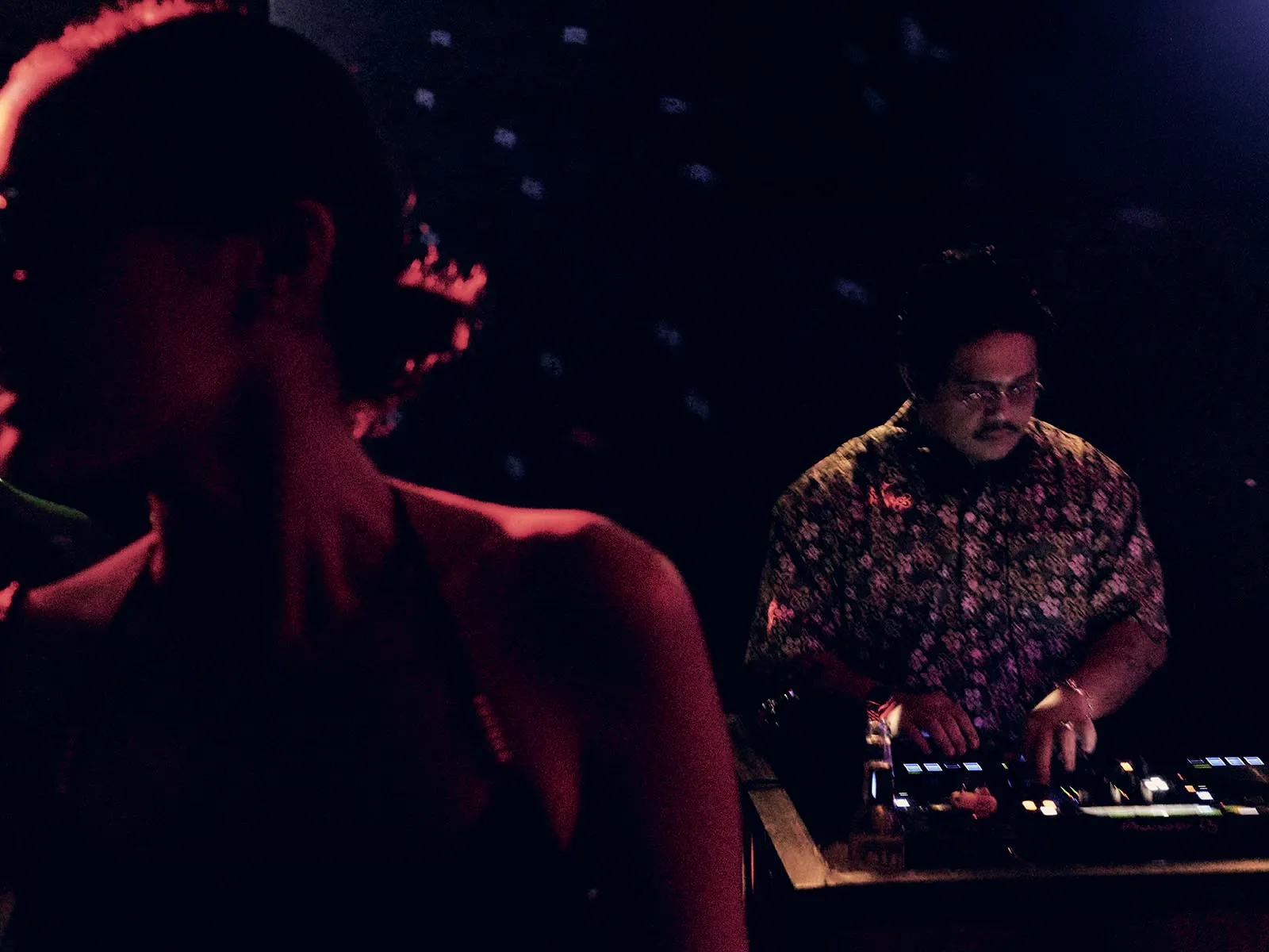 DJ in a patterned shirt mixes on turntables in a dark club, red lighting casting a moody glow; a silhouette of a person is in the foreground left.