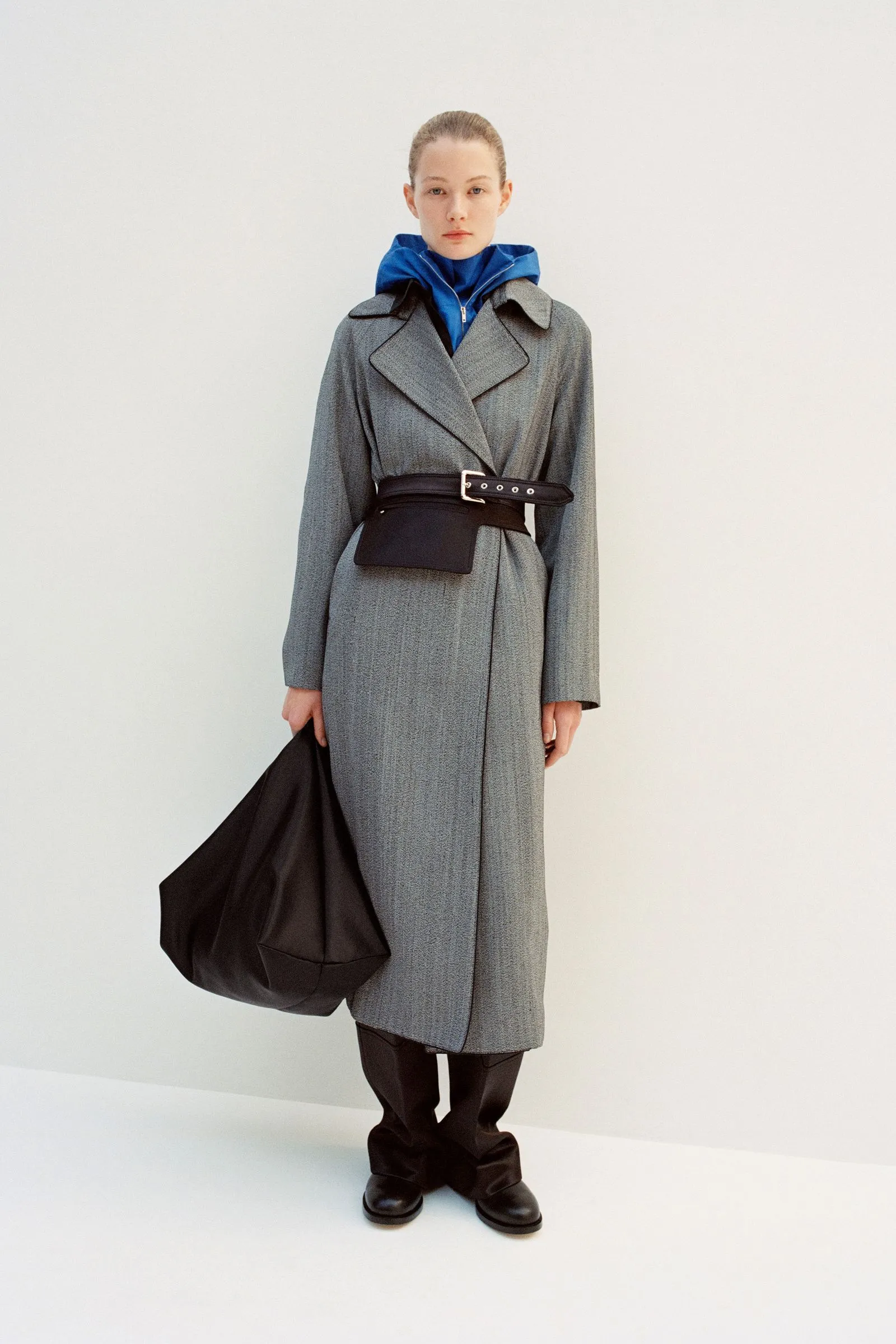Person wearing a gray long coat with a blue hooded top, cinched at the waist with a wide black belt, holding a large black bag.