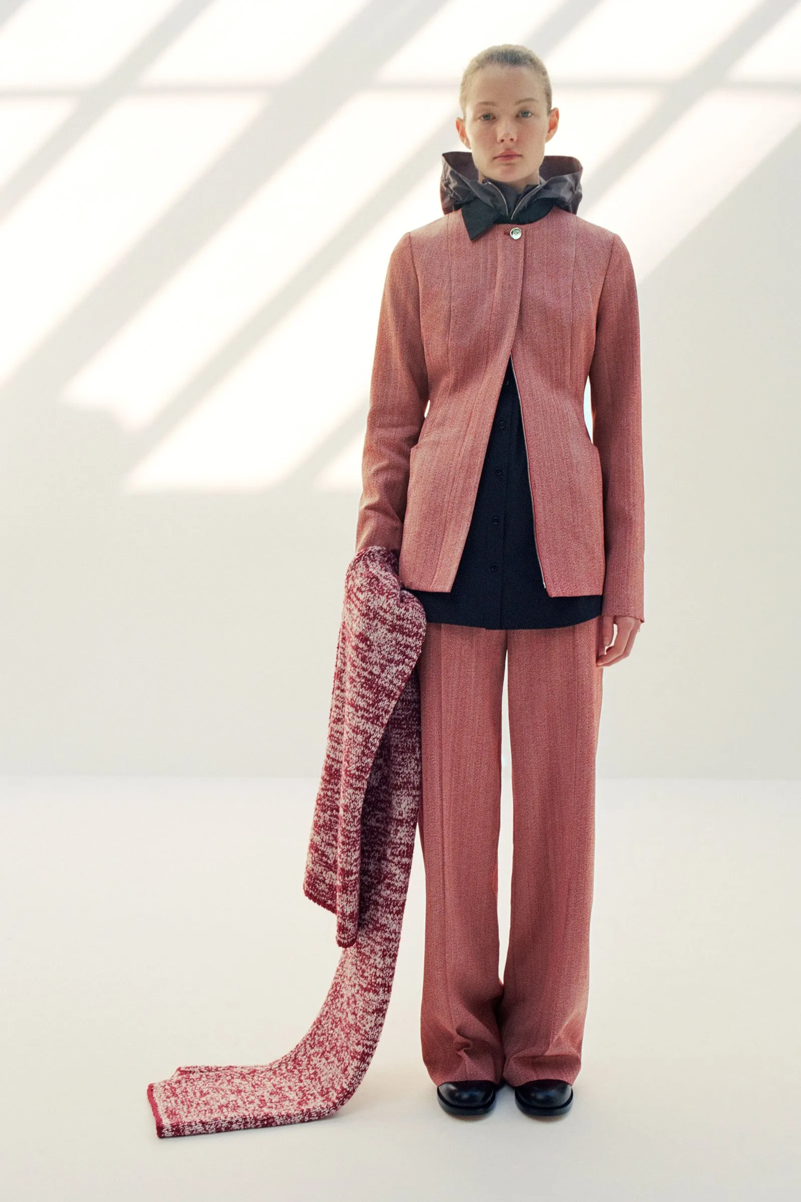 Model in a pink tailored suit with a black inner top and oversized black neck scarf, a long pink-and-white knitted scarf drapes to the floor beside her.