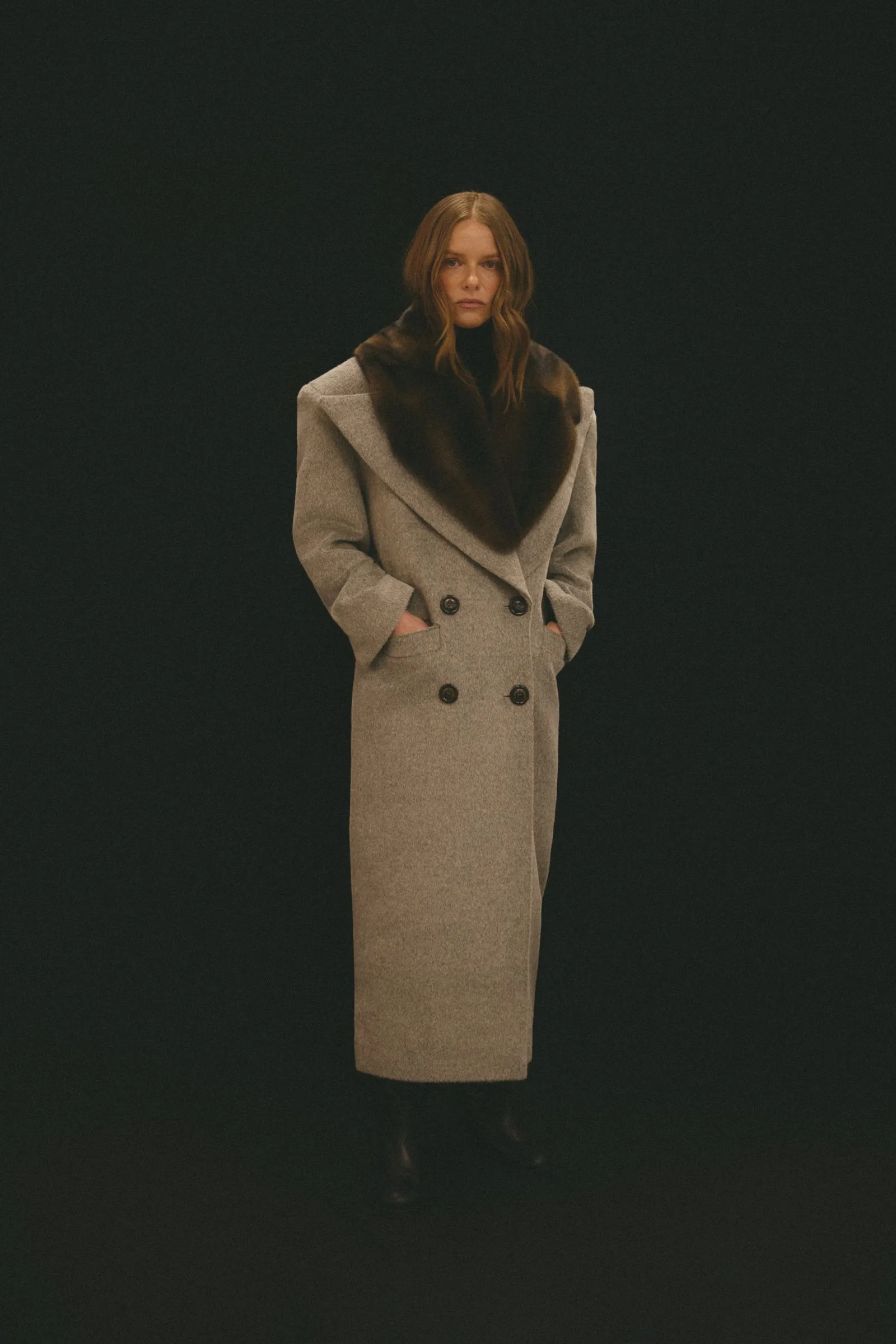 Model wearing a light gray wool coat with a dark fur collar, standing against a black backdrop.