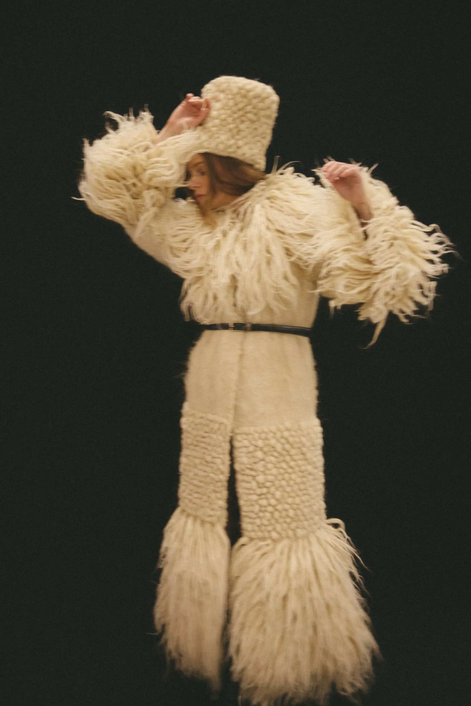 Performer wearing a long cream shaggy coat and tall furry hat on stage, arms raised in a dance pose against a black background.
