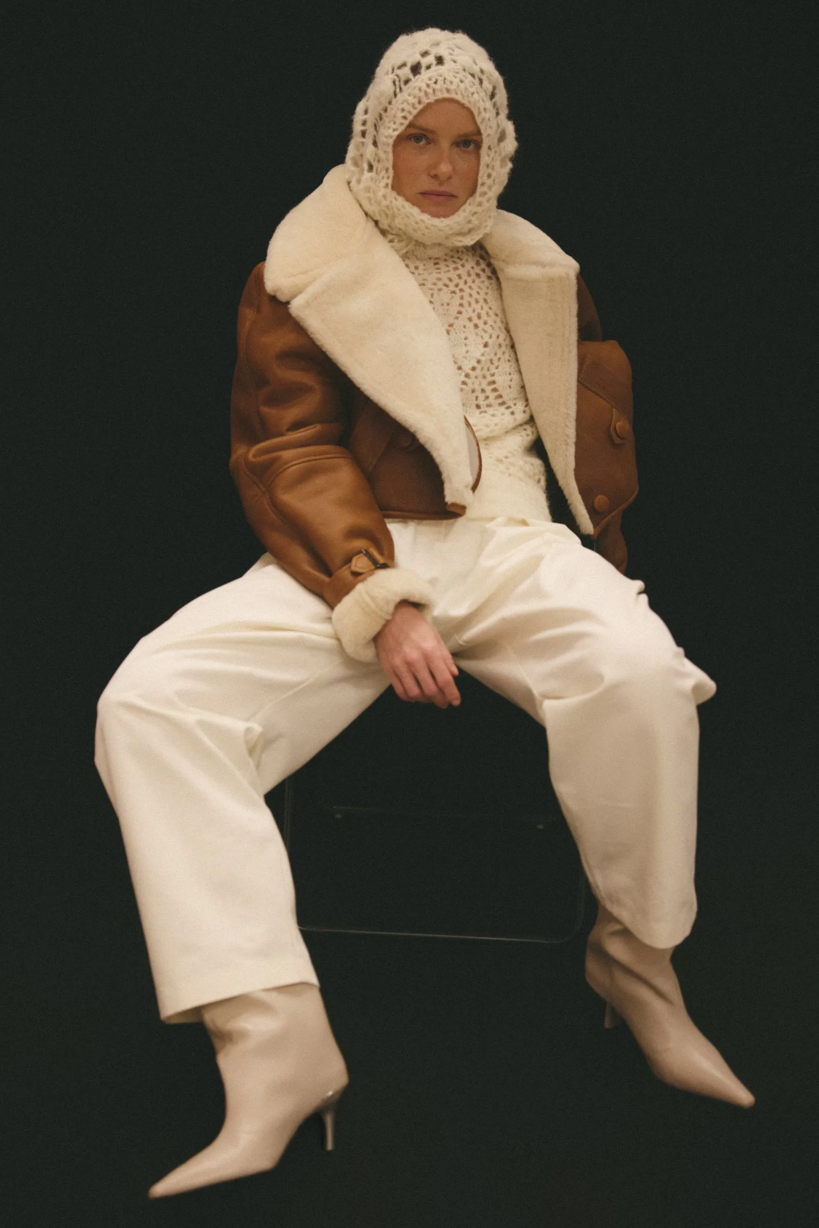 Model seated against a dark background, wearing a cream knit hood, cream crocheted top, tan shearling jacket, and white wide-leg pants with heeled boots.