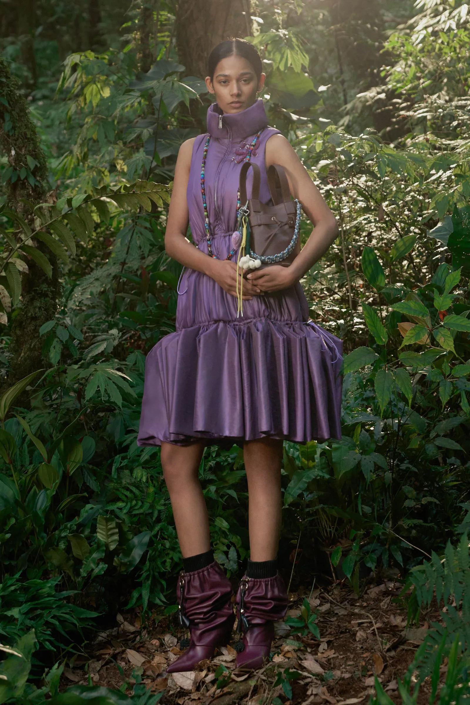 Person standing in a dense forest wearing a purple sleeveless dress, chunky boots, and colorful beaded necklaces.