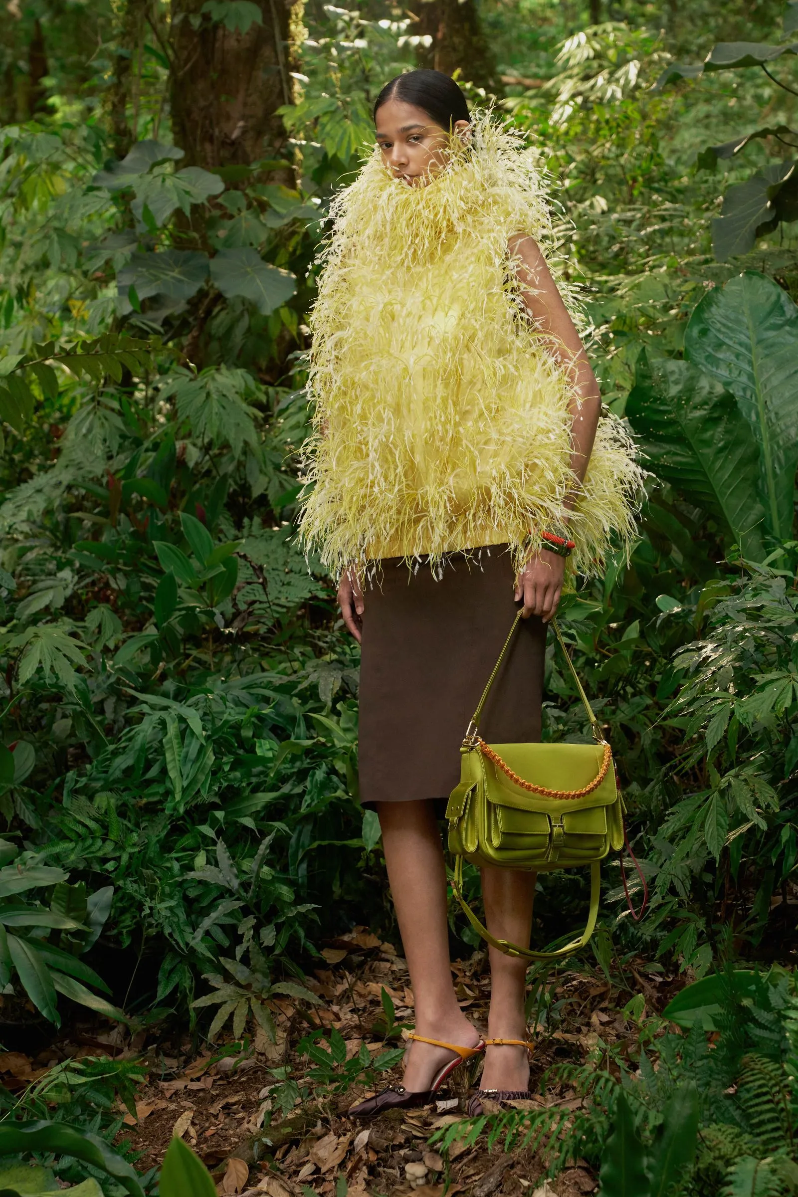 Person standing in a lush green outdoor setting wearing a bright, feathered yellow top and a brown skirt, carrying a green handbag with a yellow strap/orange chain.