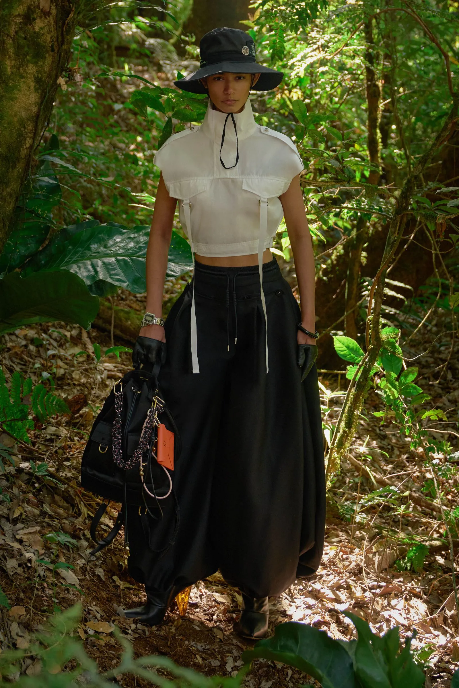 Person in a forest wearing a white cropped top, black wide pants, a black hat, gloves, and carrying a black bag.
