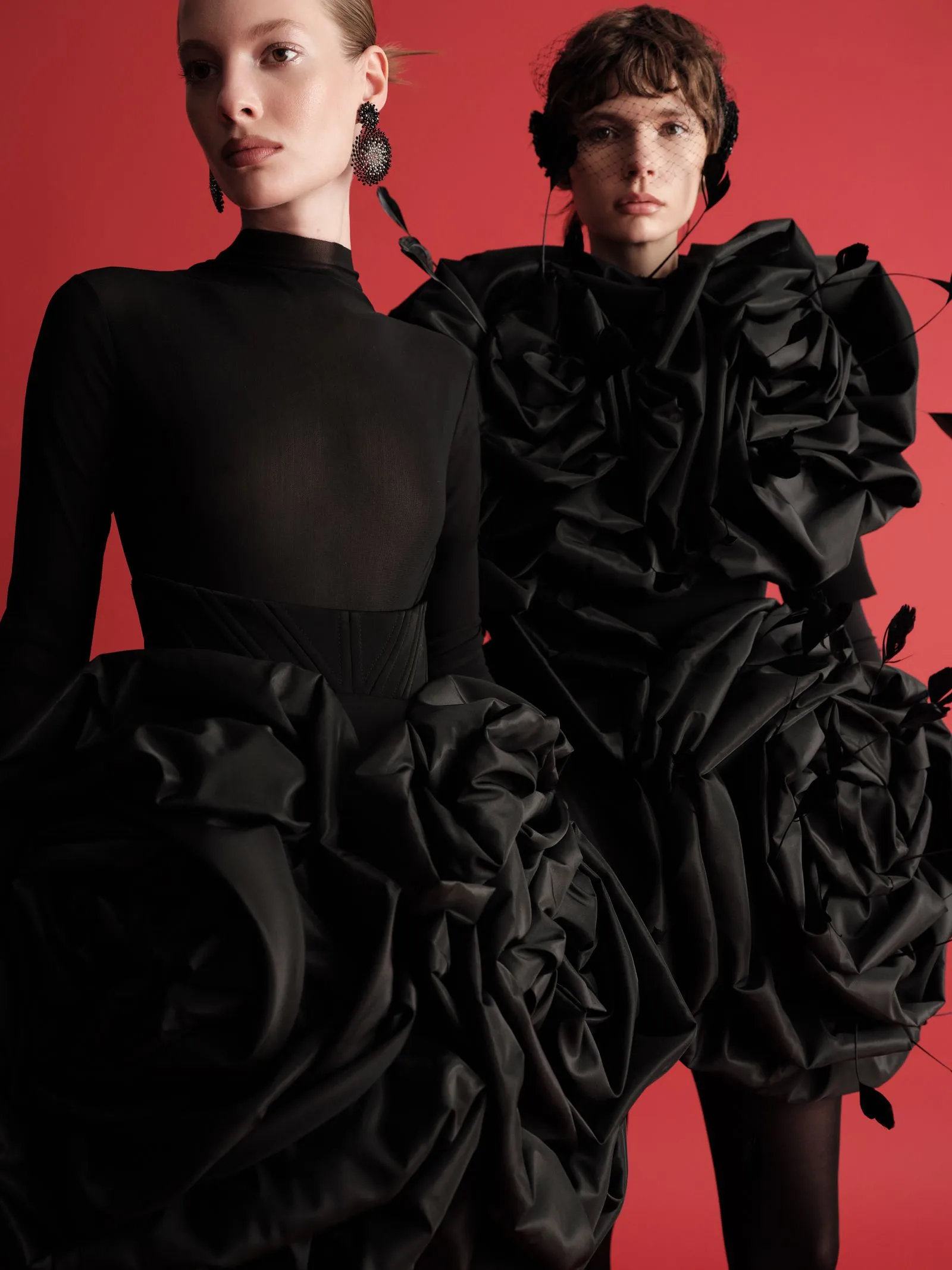 Two models in dramatic black outfits pose against a red backdrop; one in a sleek dress with statement earrings, the other in voluminous ruffled fabric and a veil-like headpiece.