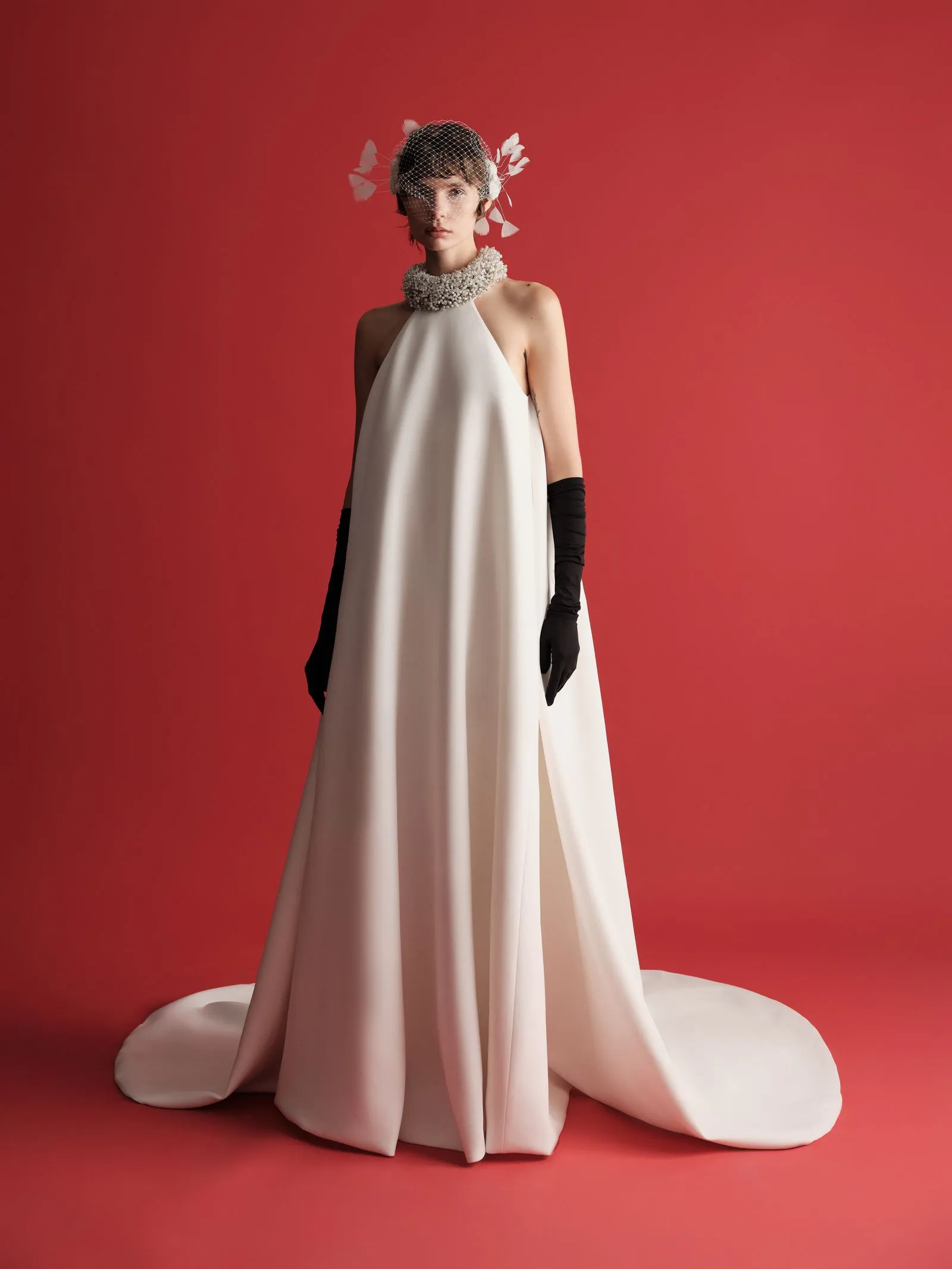 Fashion portrait of a woman in an ivory halter gown with a pearl-embellished collar, veil, and long black gloves against a solid red background.