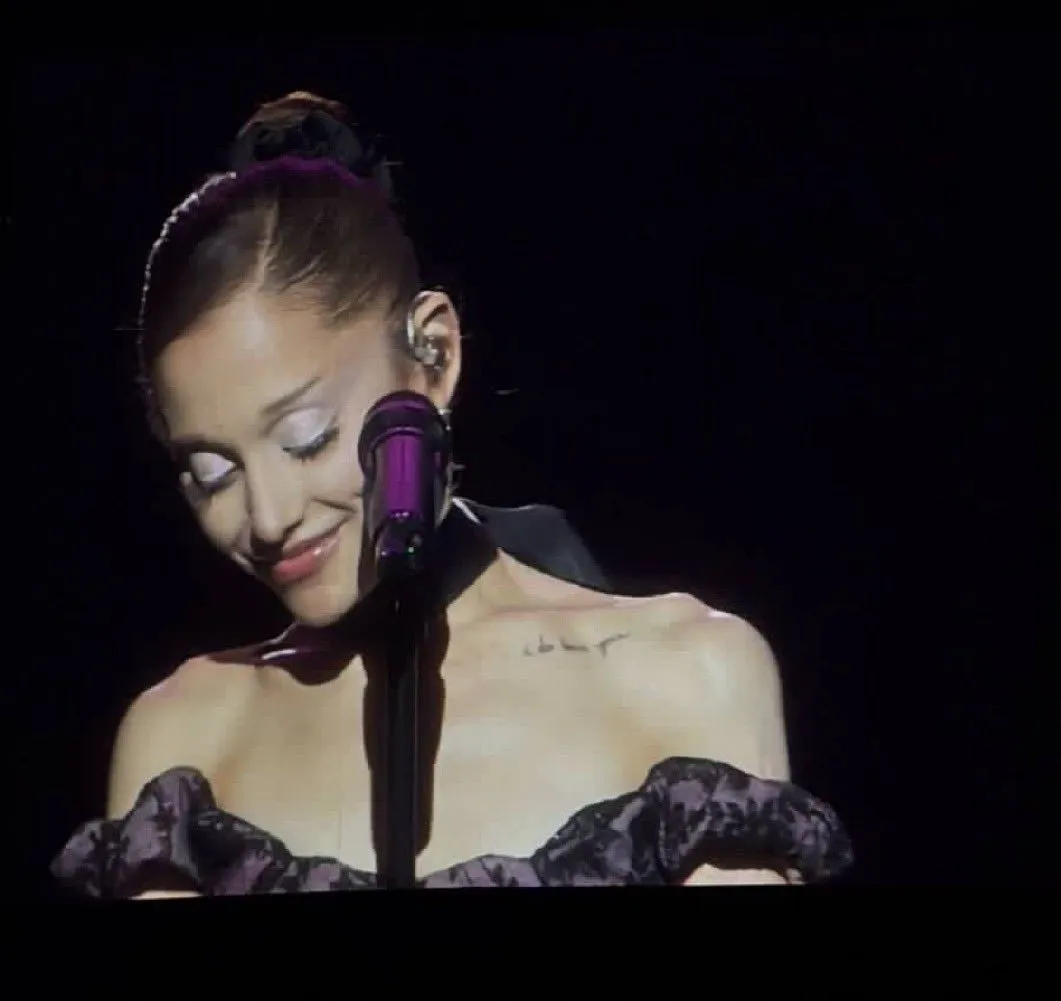 Female singer on stage with a purple microphone, wearing an off-shoulder ruffled dress, eyes closed in a moment of emotion.