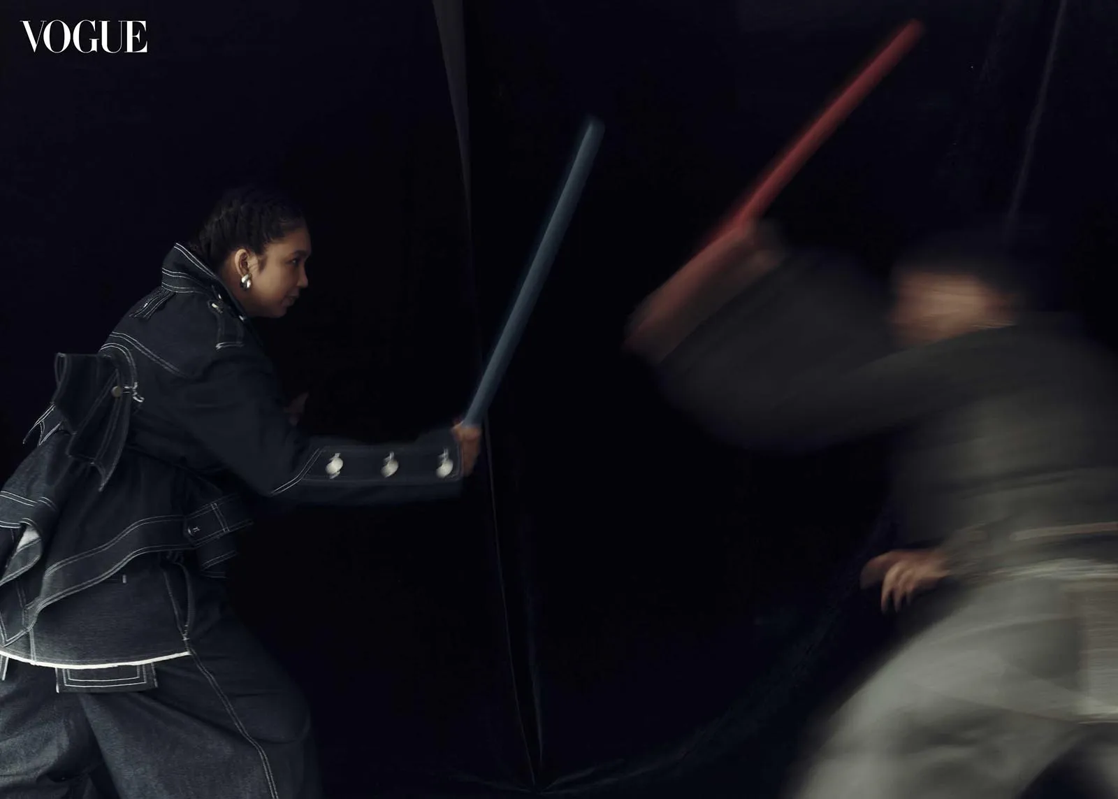 Model in oversized dark denim, reaching with a blue baton toward a blurred figure against a dark background; Vogue logo in the top-left corner.