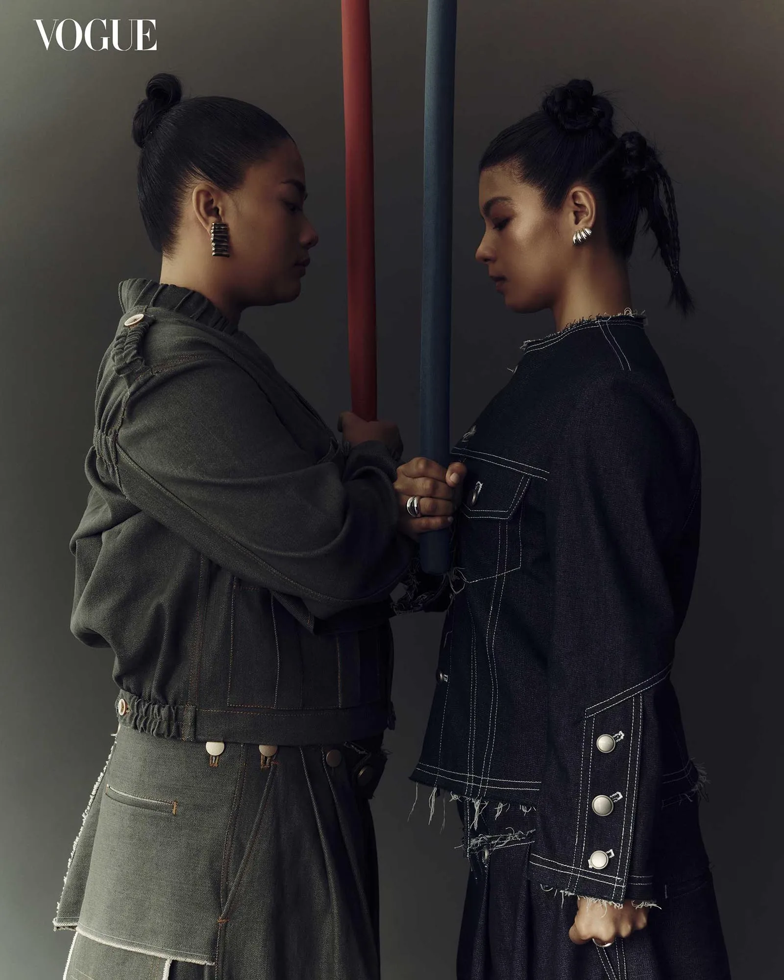 Two women in denim jackets face each other, gripping red and blue poles against a dark studio backdrop; Vogue logo in top-left corner.