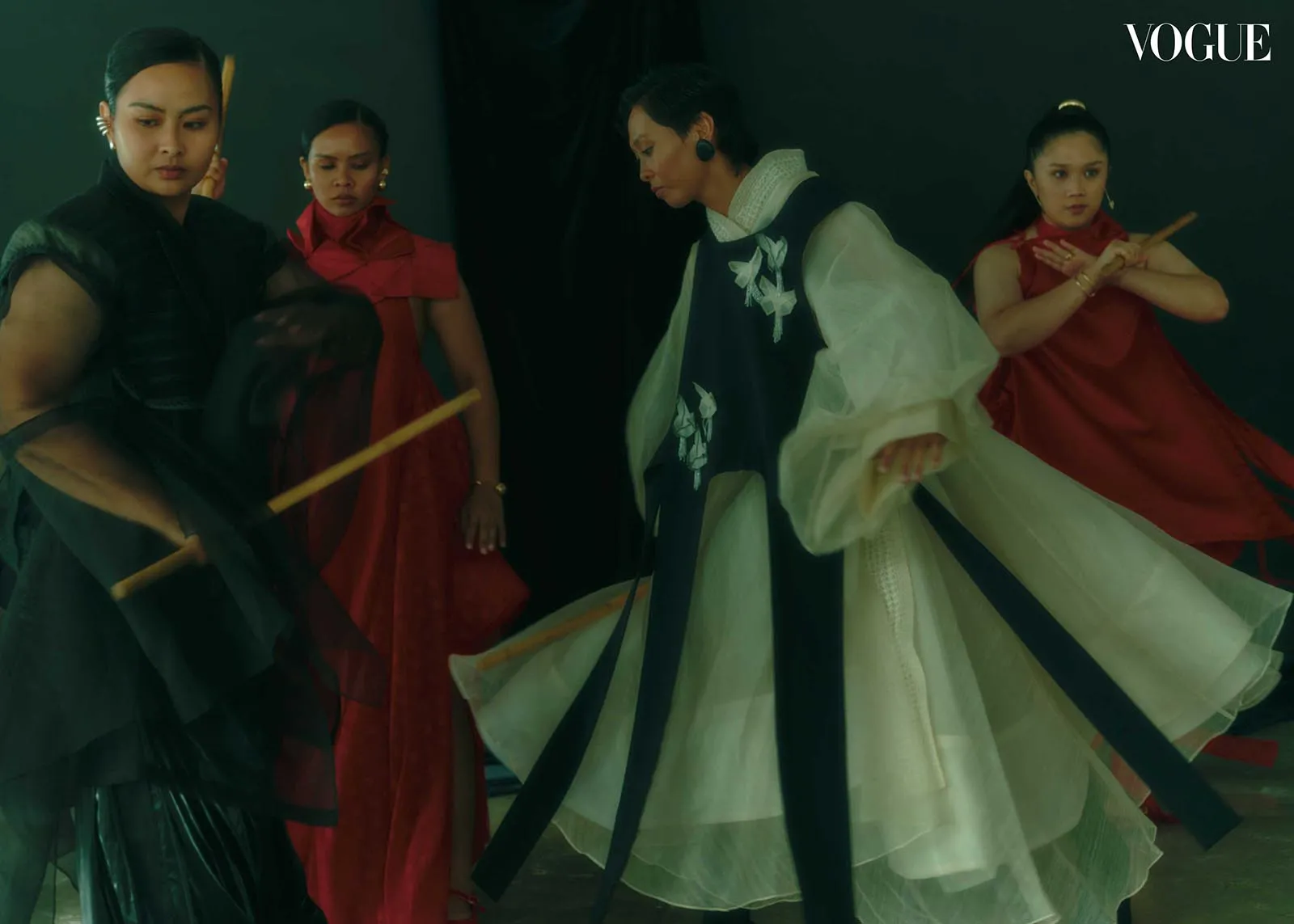 Editorial fashion shot: models in red and dark gowns swirl with flowing fabrics on a dark studio backdrop, Vogue logo visible.