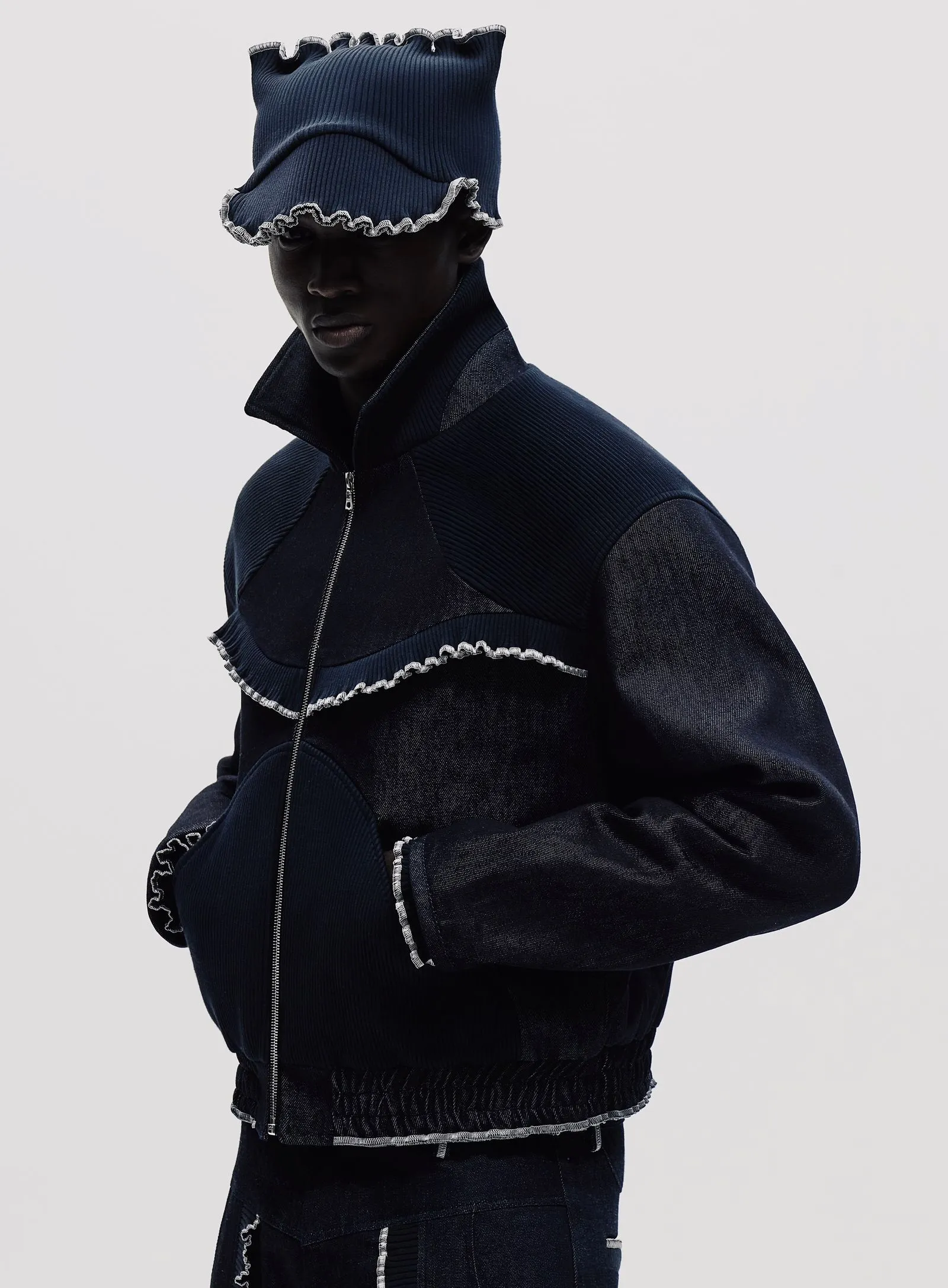 Model wearing a navy denim jacket with a matching ruffled hood and white-stitched edges, standing against a light background.