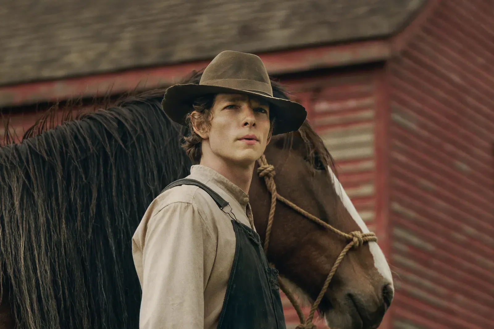 Young man wearing a brown hat and overalls stands beside a dark horse tied with rope, in front of a weathered barn.