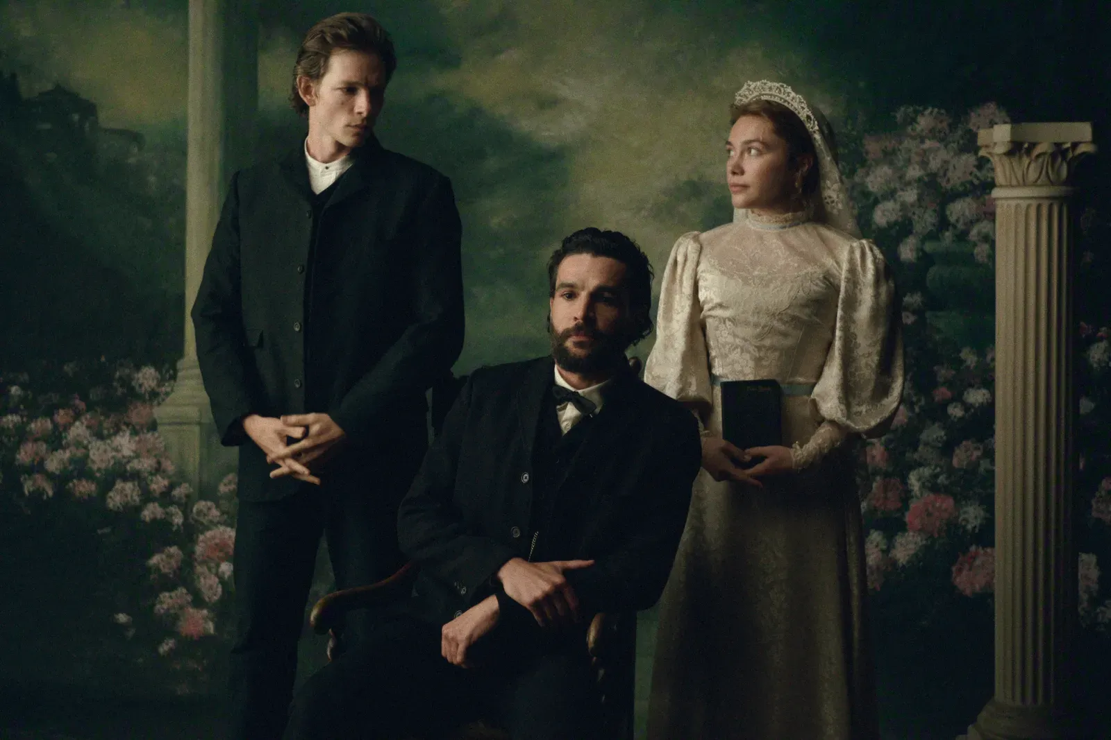 Bearded man in a black suit seated center, flanked by two formally dressed companions in a floral garden setting with a column on the right.