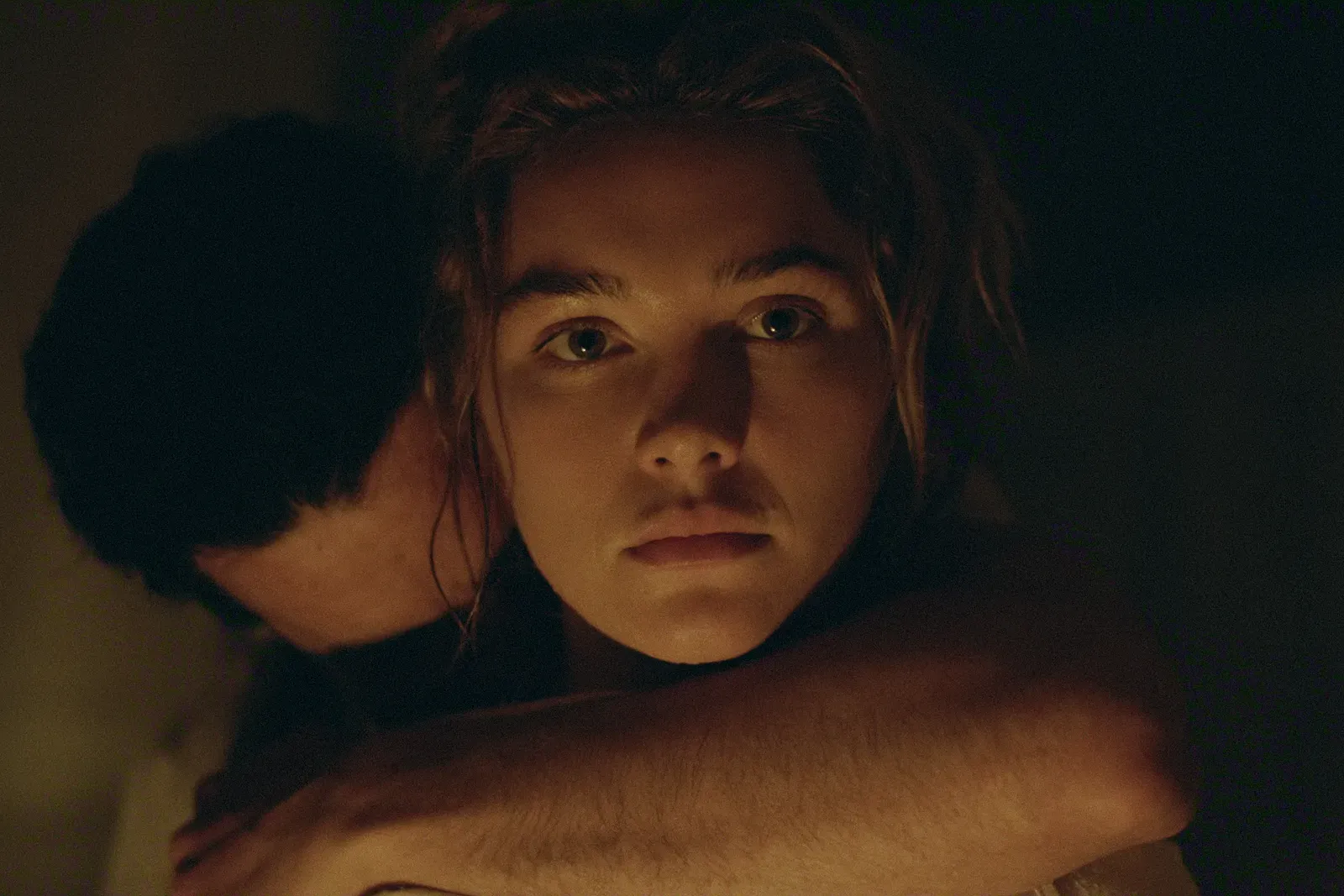 Close-up of a young woman’s face in warm, low light, looking upward with a calm, wary expression while being held from behind by another person.