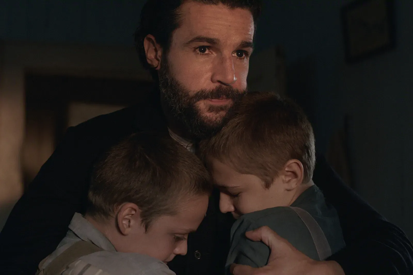 A bearded man comforting two children in a close hug in a dimly lit room.