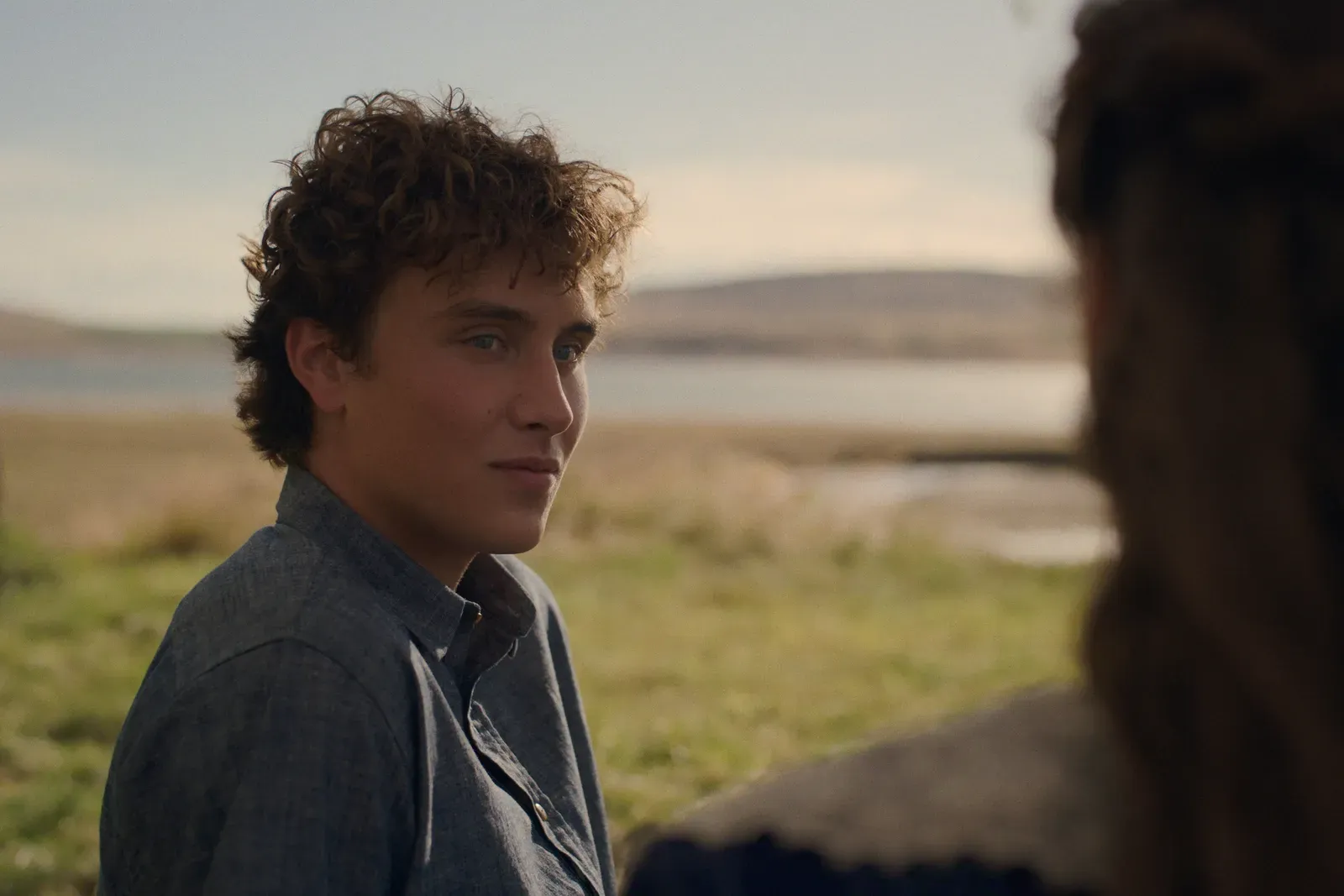 Young person with curly hair wearing a gray shirt sits outdoors by a lake, looking toward someone off camera.
