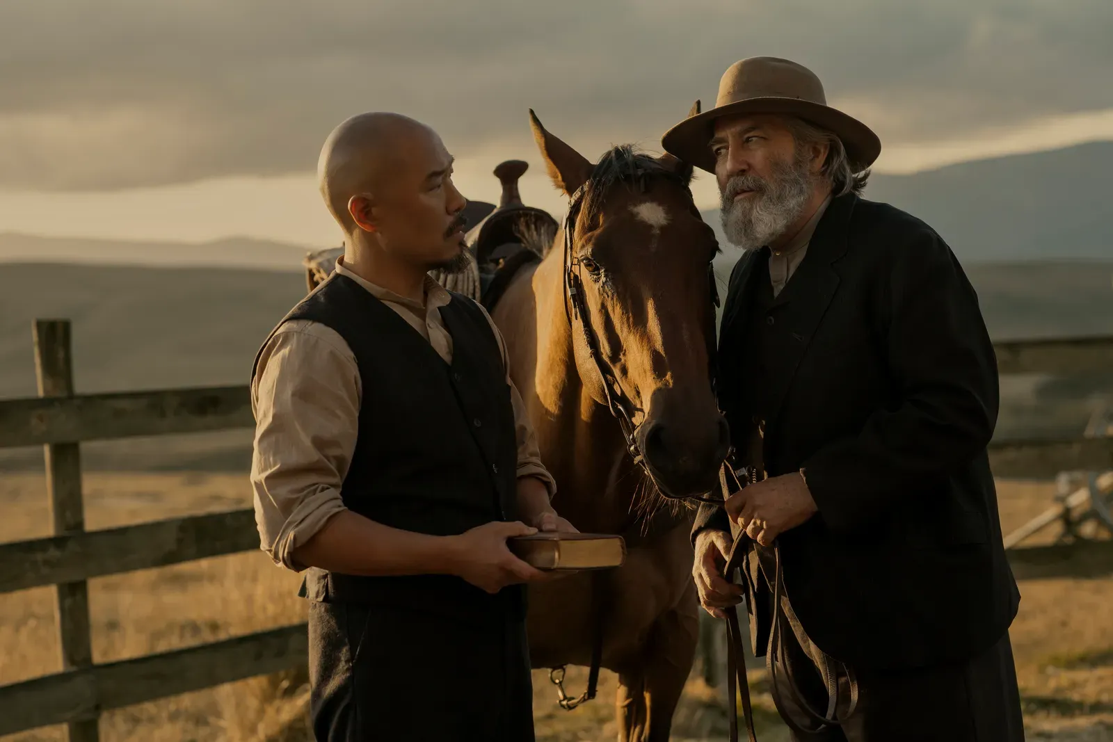 Two men in period clothing stand by a horse in a fenced, rural setting at sunset; one holds a book while the other wears a wide-brimmed hat and speaks to the horse.