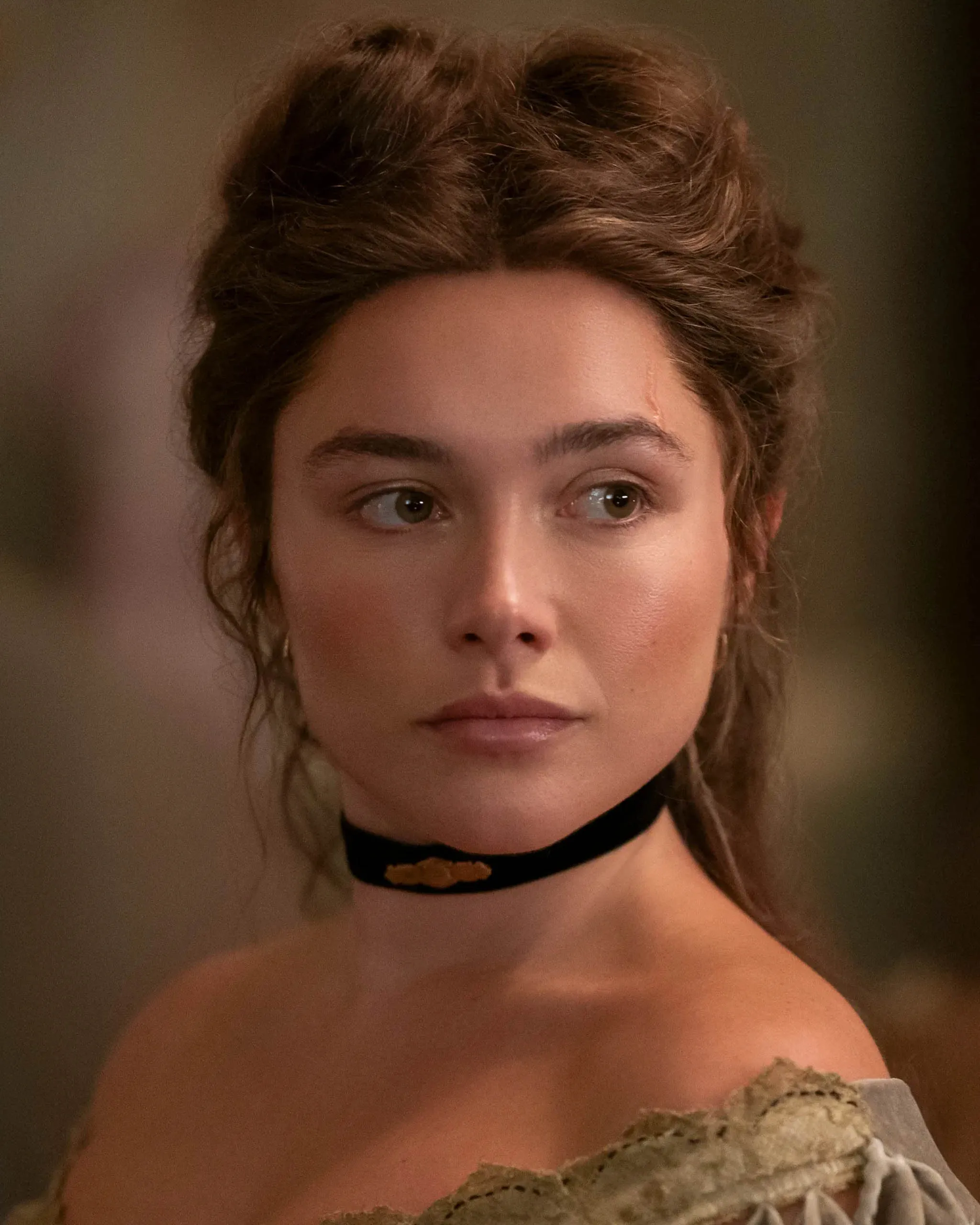 Close-up portrait of a woman with brown hair styled up, wearing a black velvet choker with a gold centerpiece and a beige lace off-shoulder dress, neutral expression.