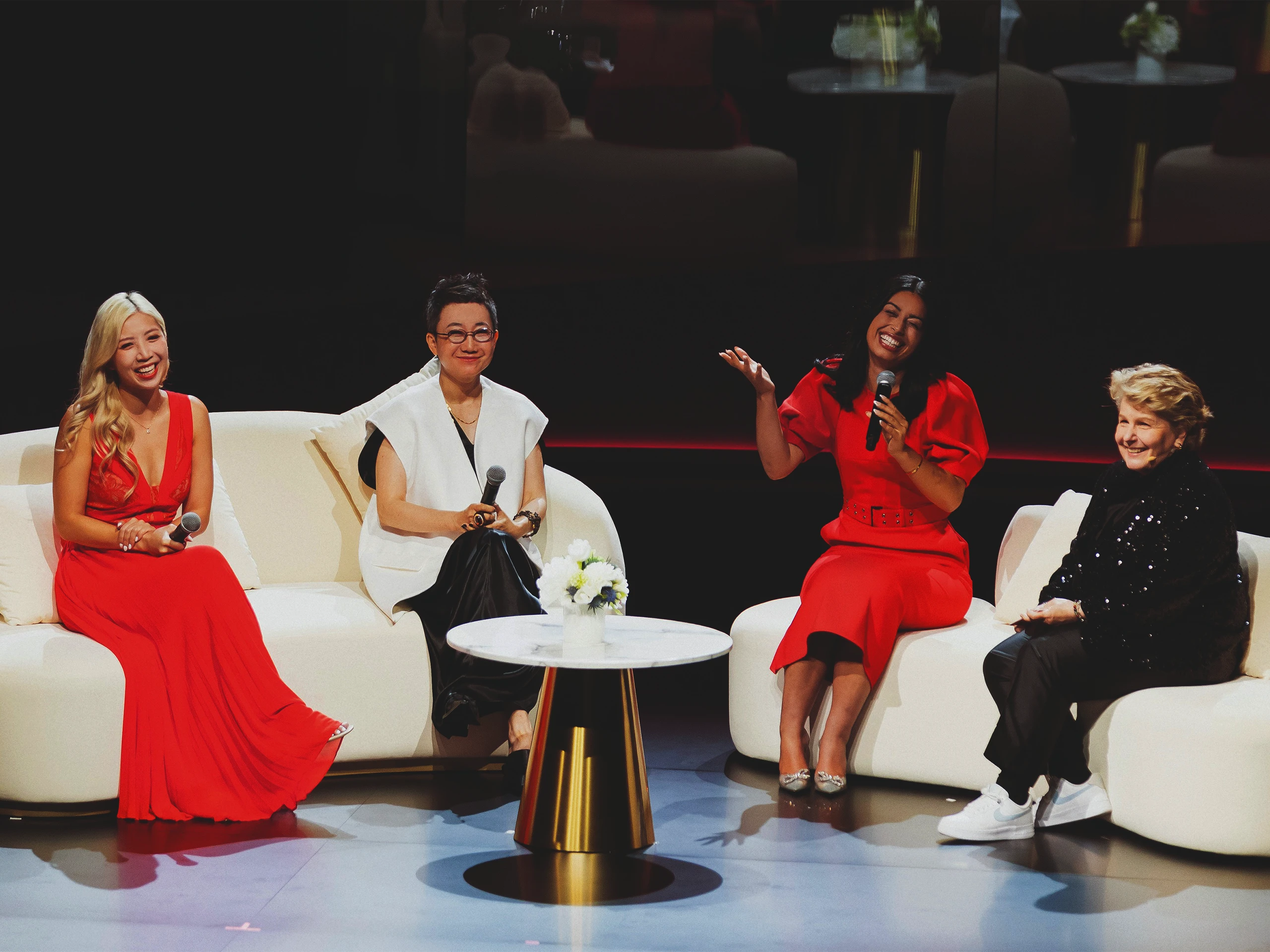 Five women in red and white outfits sit on white couches on a stage, each holding a microphone, with a small flowered table between them.