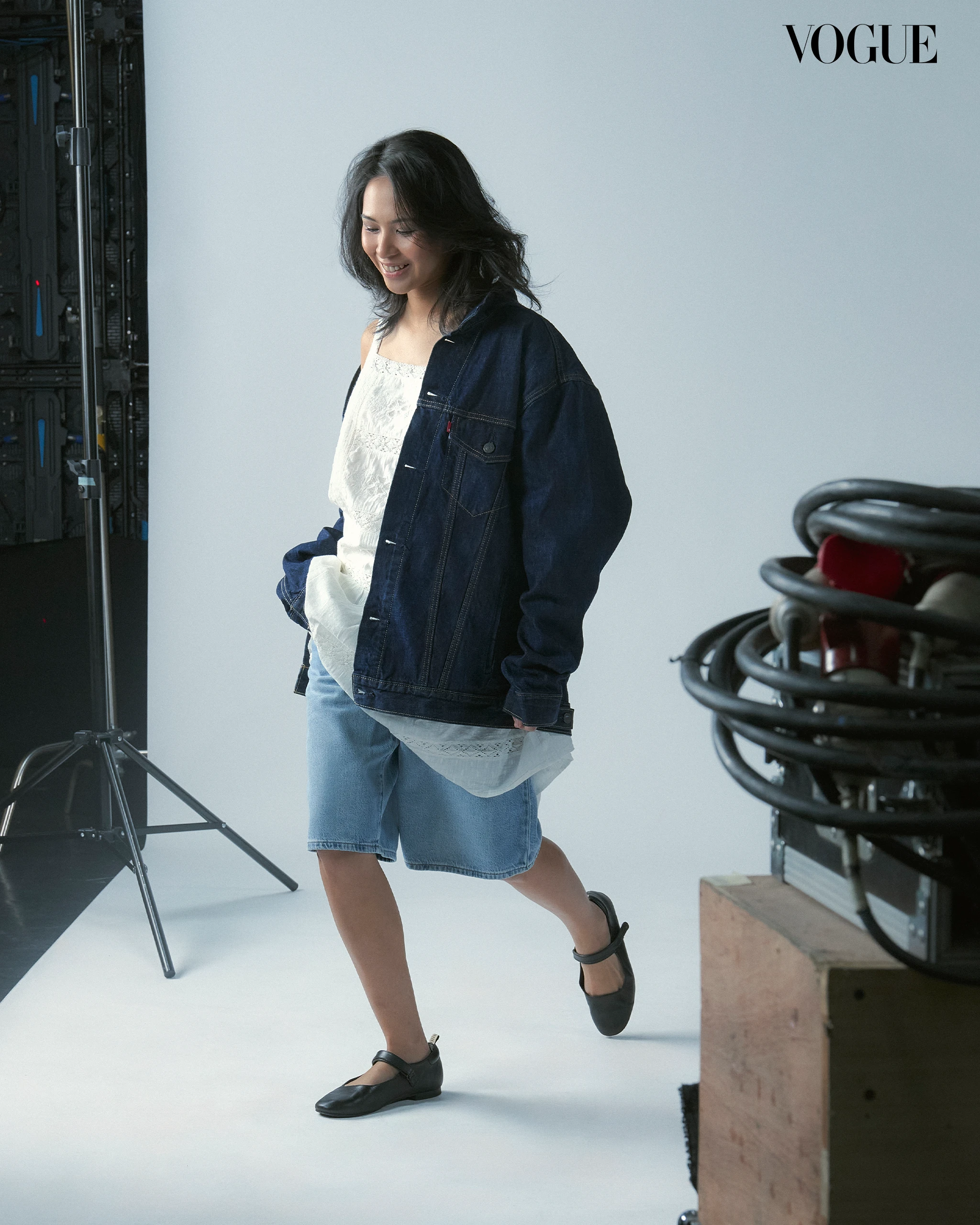Woman in denim jacket over white lace top and denim shorts walks in a studio with equipment on the left and a Vogue logo in the corner