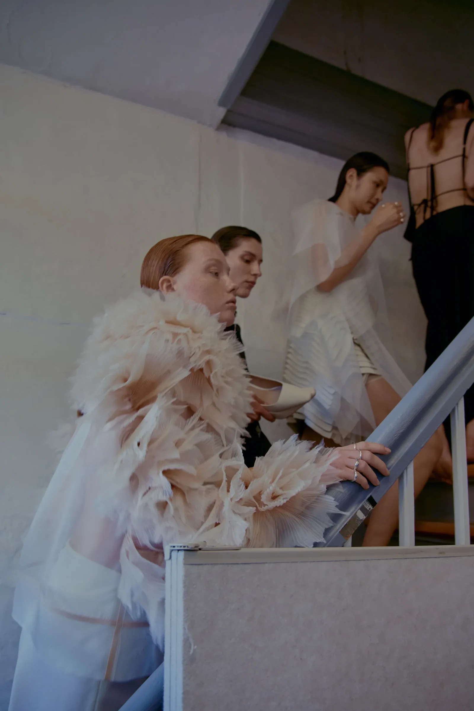 Models in white, feathered outfits descend a stairwell backstage at a fashion show, with soft lighting and a gray wall backdrop.