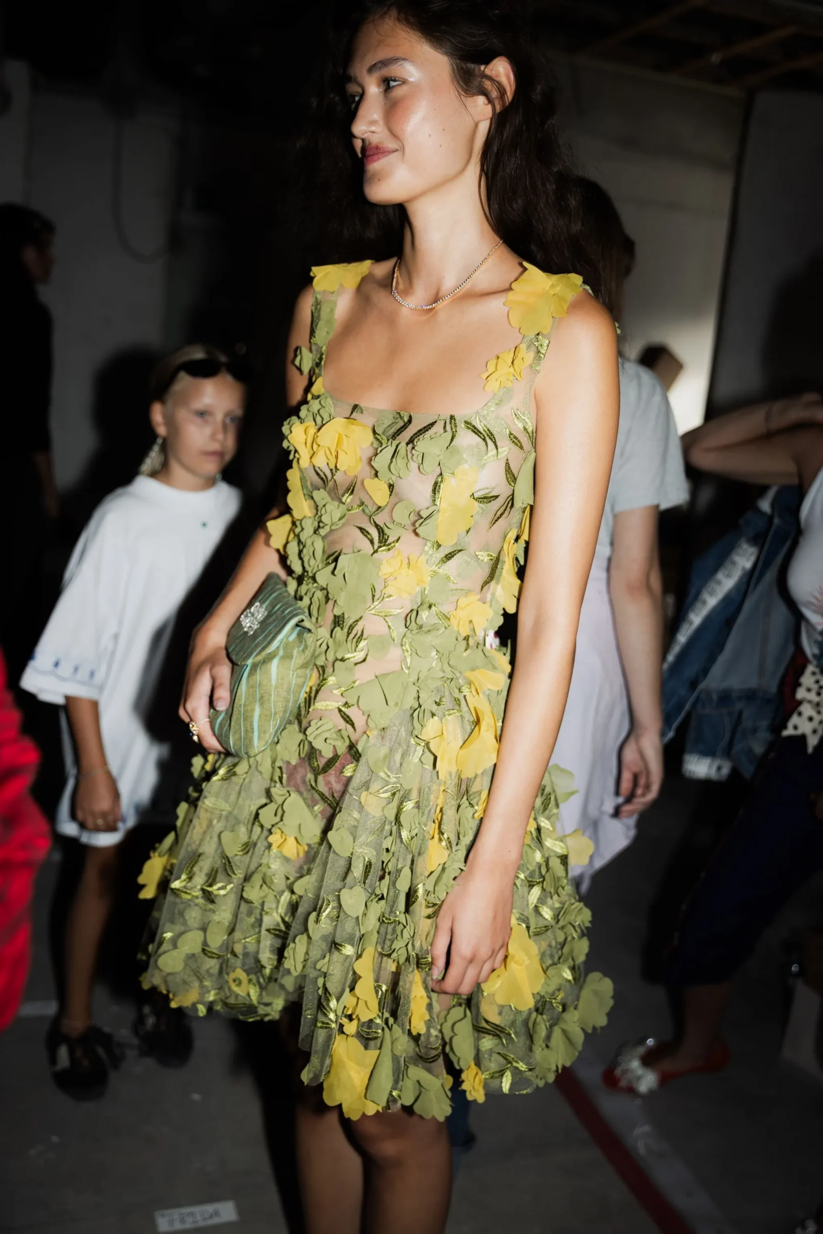Model wearing a sheer olive-green dress with yellow floral appliqués and a pearl necklace, walking backstage at a fashion show.