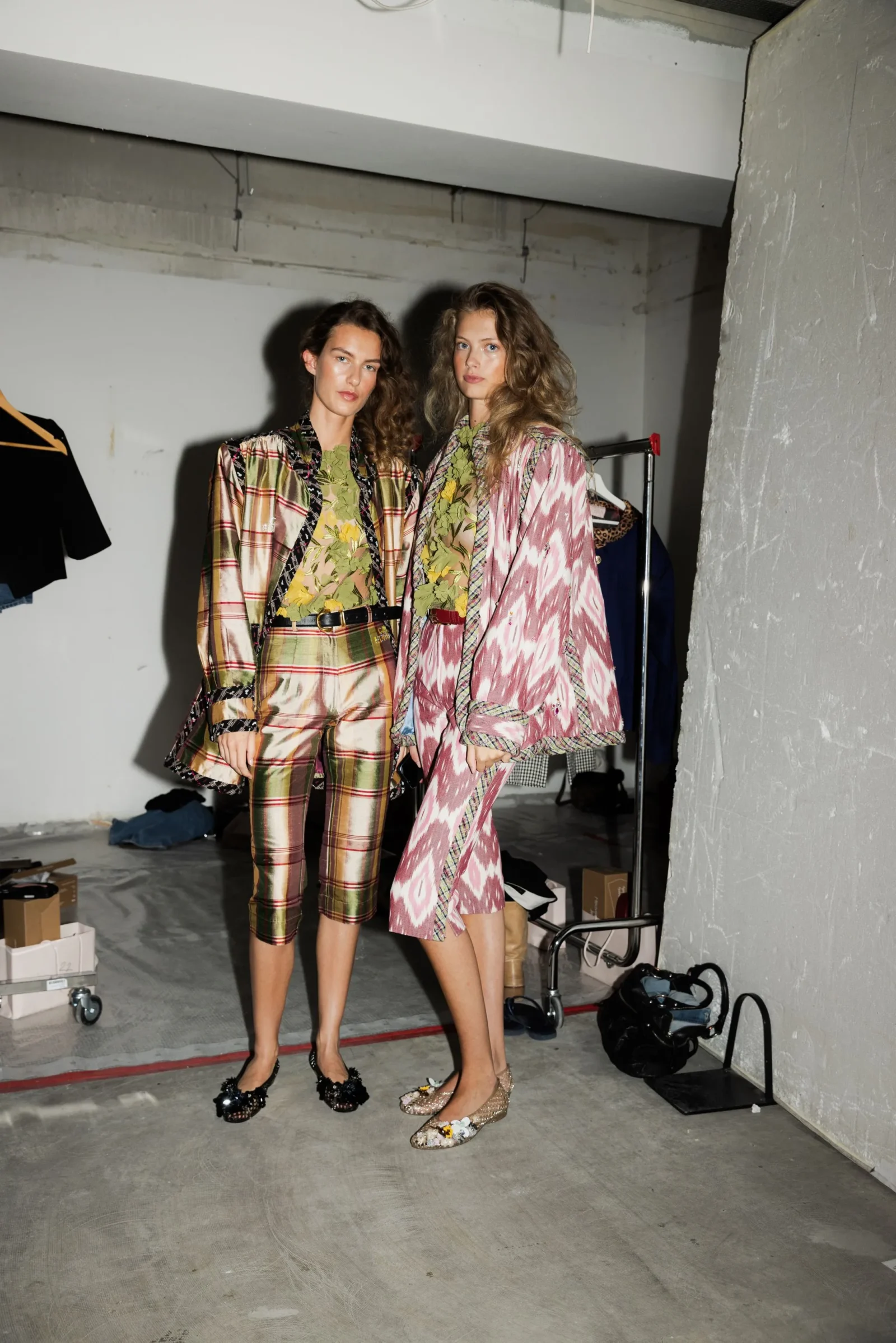 Two models pose backstage in shiny, patterned suits; one in greenish plaid and the other in pink, with a clothing rack, boxes, and shoes scattered on a concrete floor in a studio space.