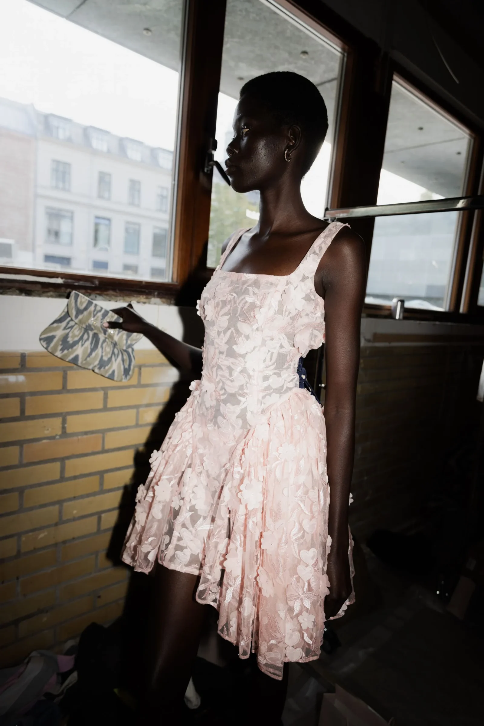 Side profile of a woman with short hair wearing a pale pink lace dress, standing by a window and holding a patterned clutch inside a dimly lit space.