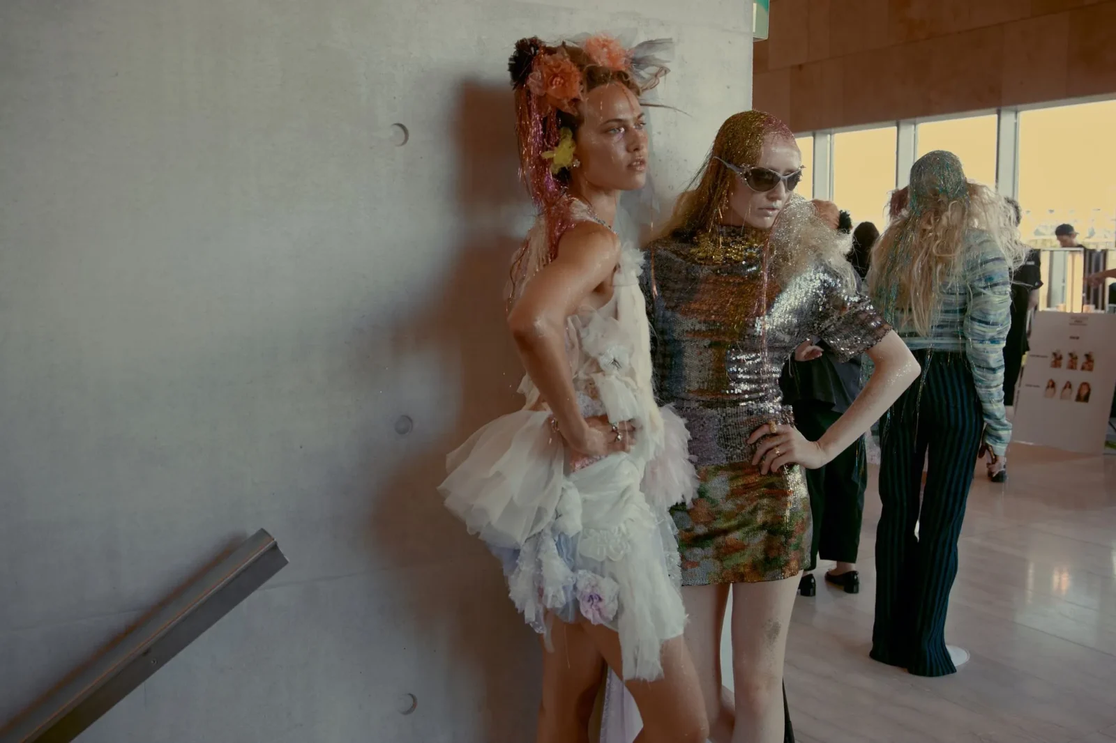 Models strike poses in glamorous, glittery outfits near a concrete wall at a fashion event.