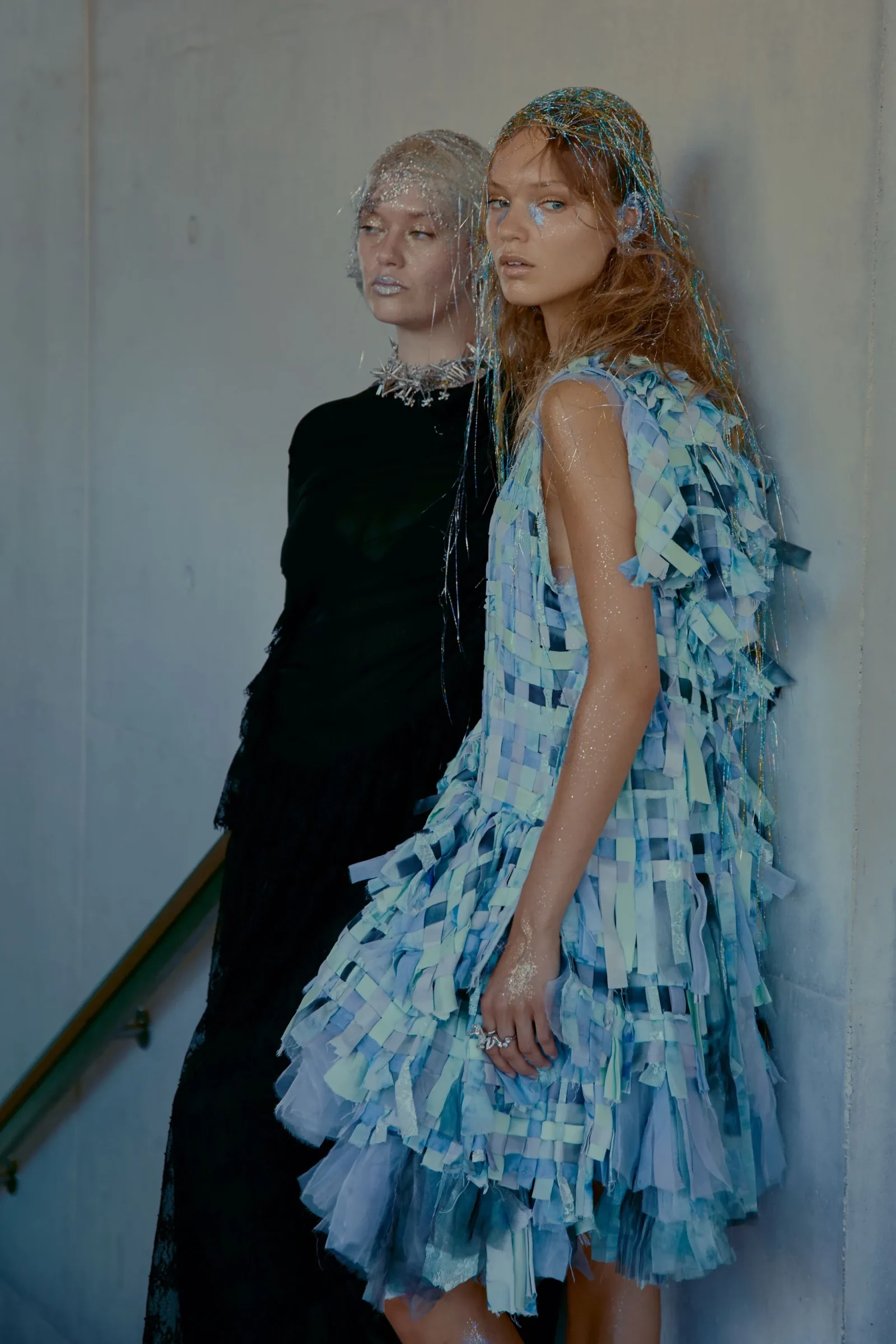 Two women posing in a stairwell wearing avant-garde dresses with blue fringe and glitter on their faces.